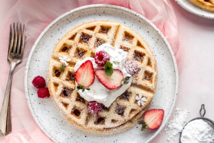 Fulffy yeast Belgian waffle on white plate topped with whipped cream and strawberries. The plate is sitting on a light pink background and surrounded by a bowl of strawberries, powdered sugar in a sifter, and a pink glass of milk