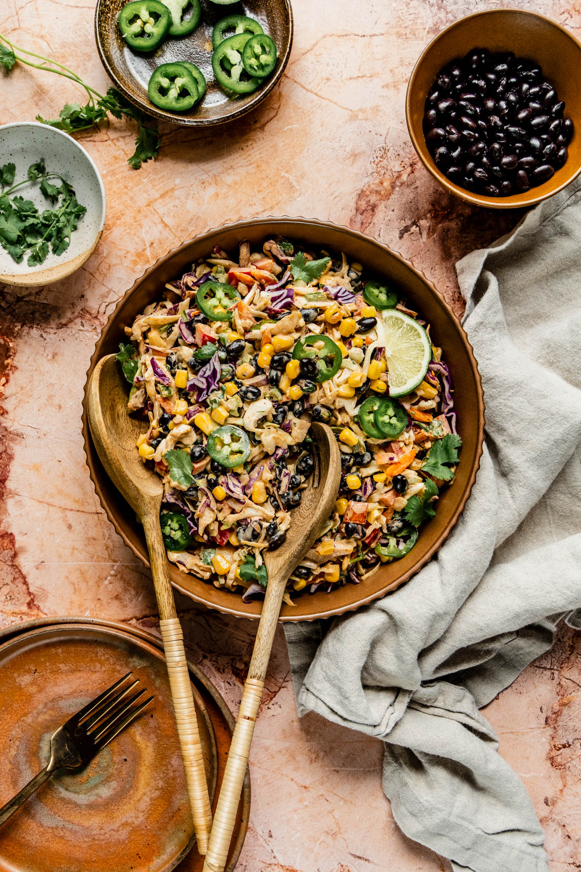 summer slaw in brown bowl on brown marble background with salad tongs in bowl. Also on the counter is a bowl of black beans, a dish with sliced jalapeno and a bowl of fresh cilanto.