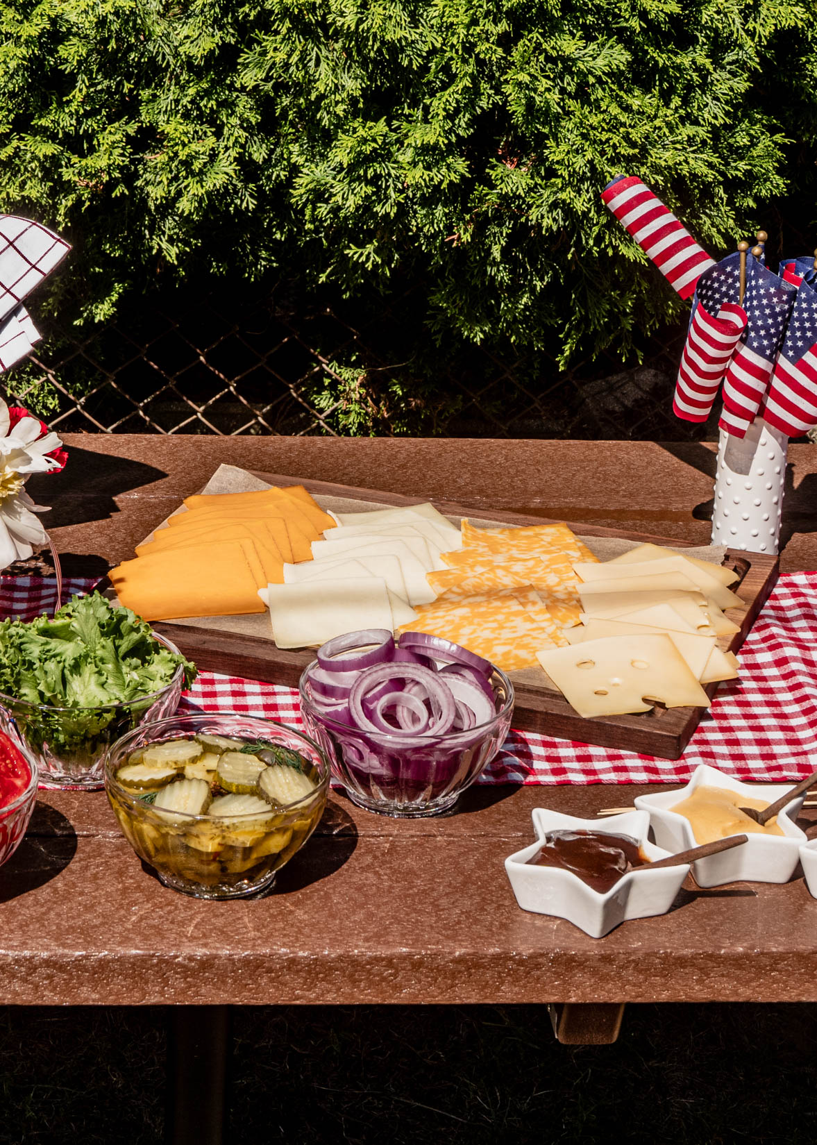 A festive July 4th Burger Bar topping station featuring sliced cheeses, red onions, pickles, lettuce, and patriotic star-shaped ramekins filled with burger sauces, styled on a red gingham tablecloth with American flags for an outdoor backyard BBQ.
