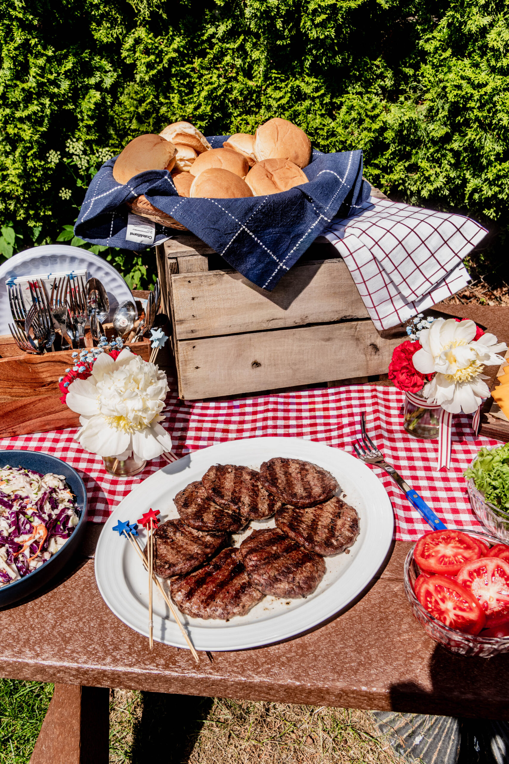 Grilled burger patties served on a white platter as part of a festive July 4th Burger Bar, styled on a red gingham tablecloth outdoors with patriotic star picks, white florals, and backyard BBQ décor.