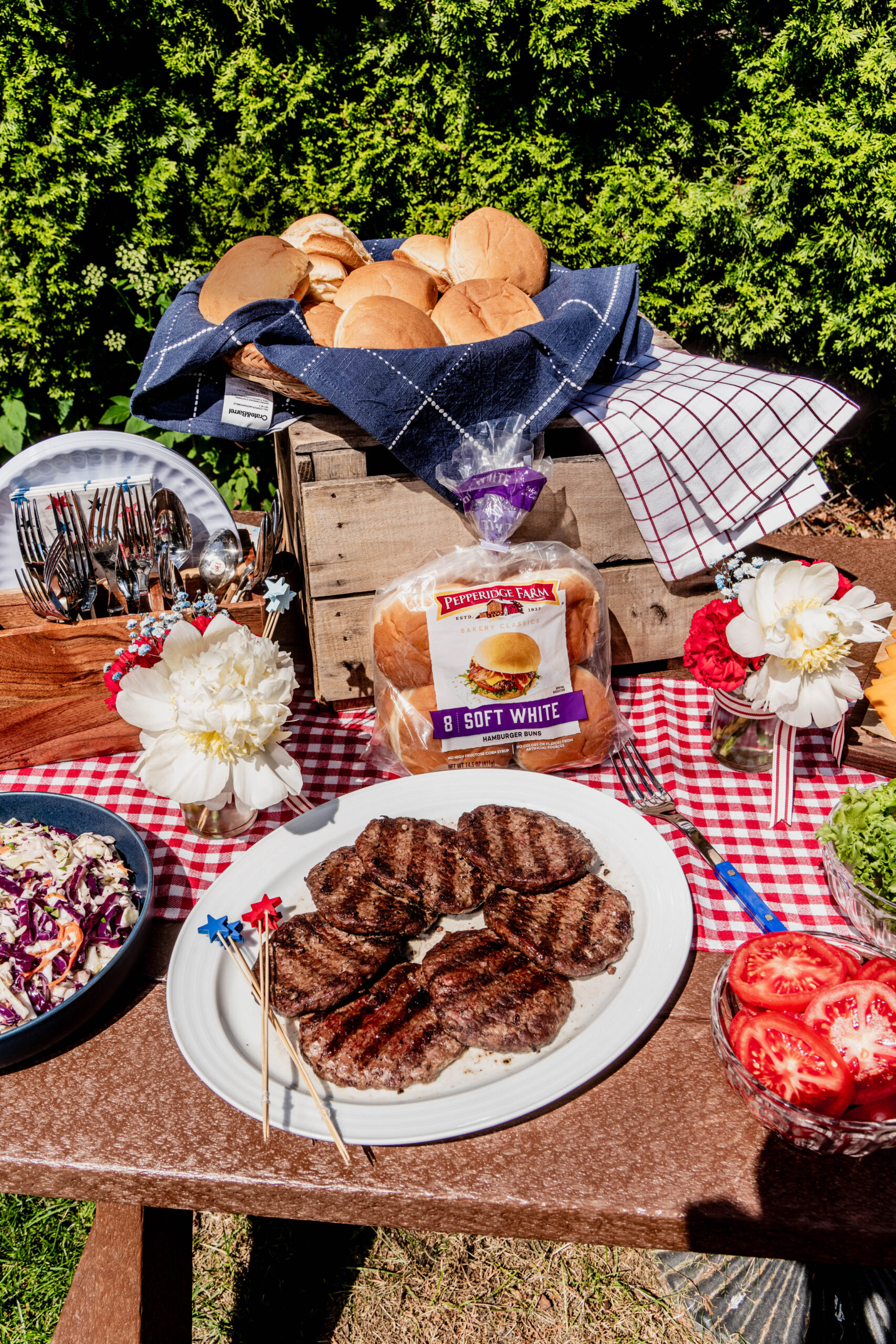 A festive outdoor July 4th Burger Bar featuring grilled burger patties, fresh hamburger buns, sliced tomatoes, lettuce, coleslaw, and patriotic décor on a red gingham tablecloth, styled with flowers, wooden crates, and classic backyard BBQ details.