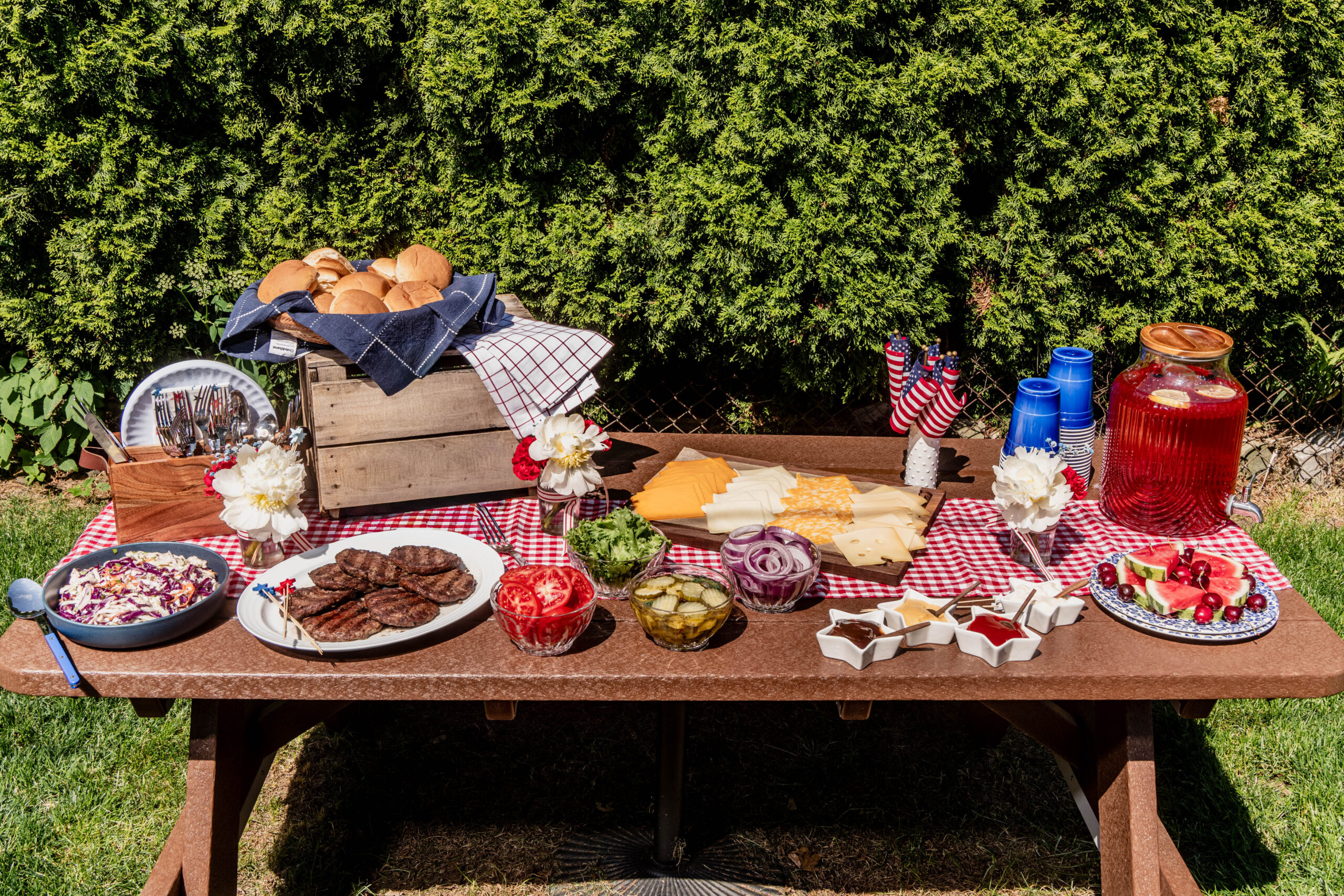 July 4th Burger Bar**Alt text:**
A festive **July 4th Burger Bar** displayed outdoors on a picnic table with a red gingham tablecloth, featuring grilled burger patties, fresh buns, sliced cheeses, tomatoes, pickles, red onions, lettuce, patriotic star-shaped ramekins filled with condiments, watermelon slices, and a red summer drink pitcher surrounded by red, white, and blue décor.