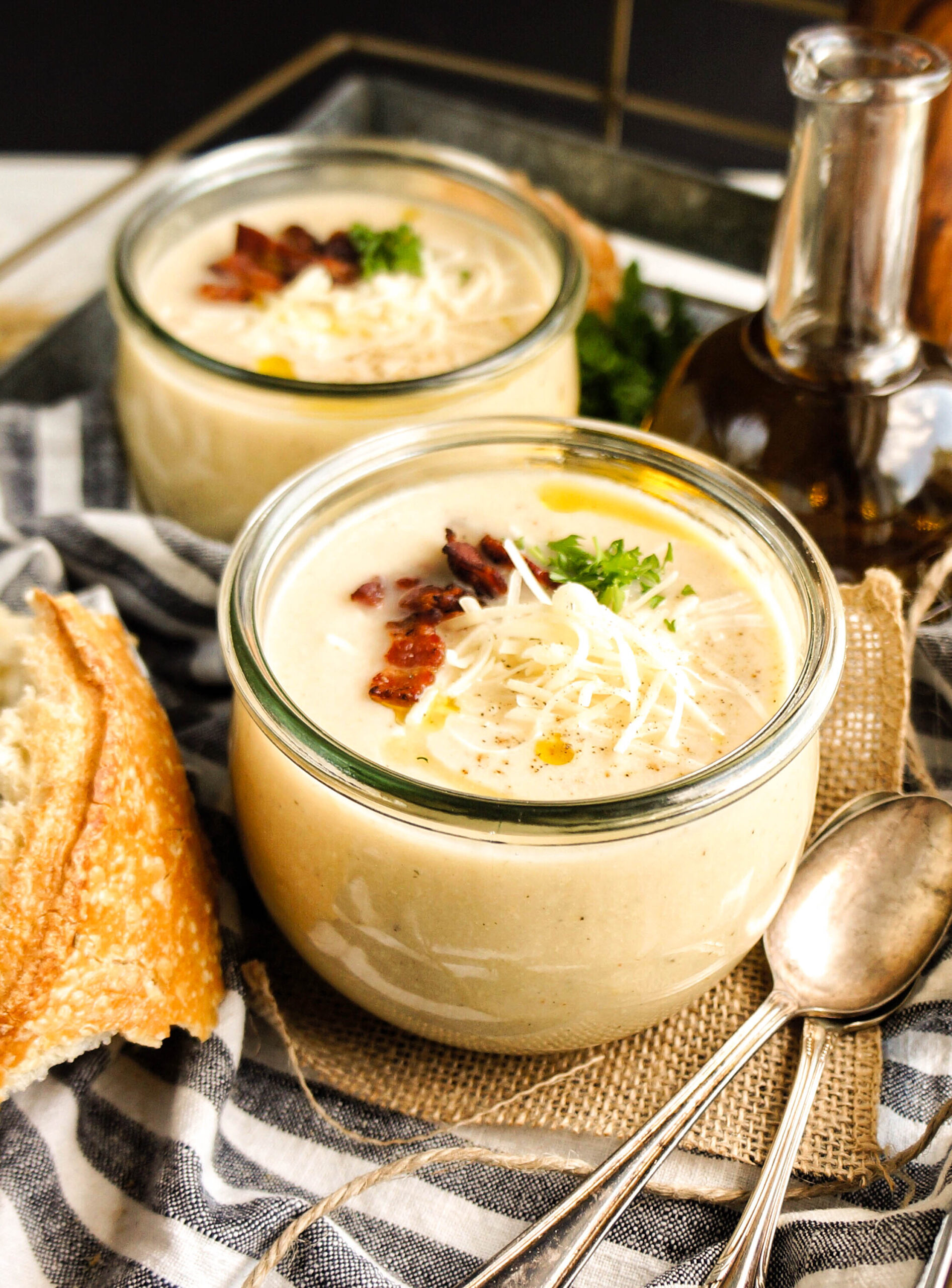 Close-up of a glass jar filled with creamy Crockpot cauliflower soup, topped with shredded cheese, crispy bacon, fresh herbs, and a drizzle of olive oil, served with a crusty baguette and spoons on a cozy neutral backdrop.