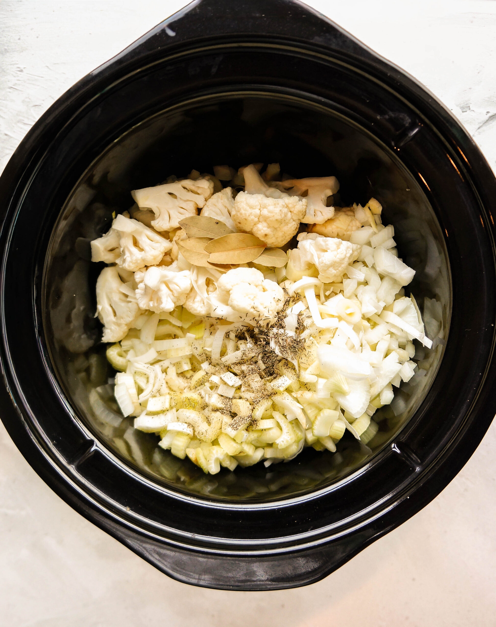 Overhead view of a slow cooker filled with raw ingredients for Crockpot cauliflower soup, including cauliflower florets, chopped leeks and onions, garlic, bay leaves, and seasonings before cooking.