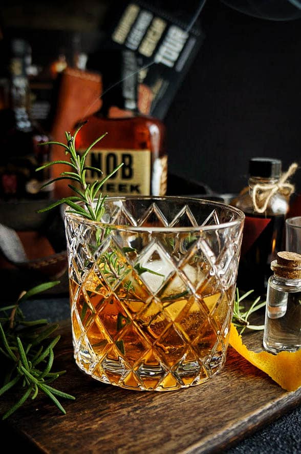 Rosemary bourbon old fashioned cocktail served over a large ice cube in a cut crystal rocks glass, garnished with a fresh rosemary sprig and orange peel on a dark wooden board.