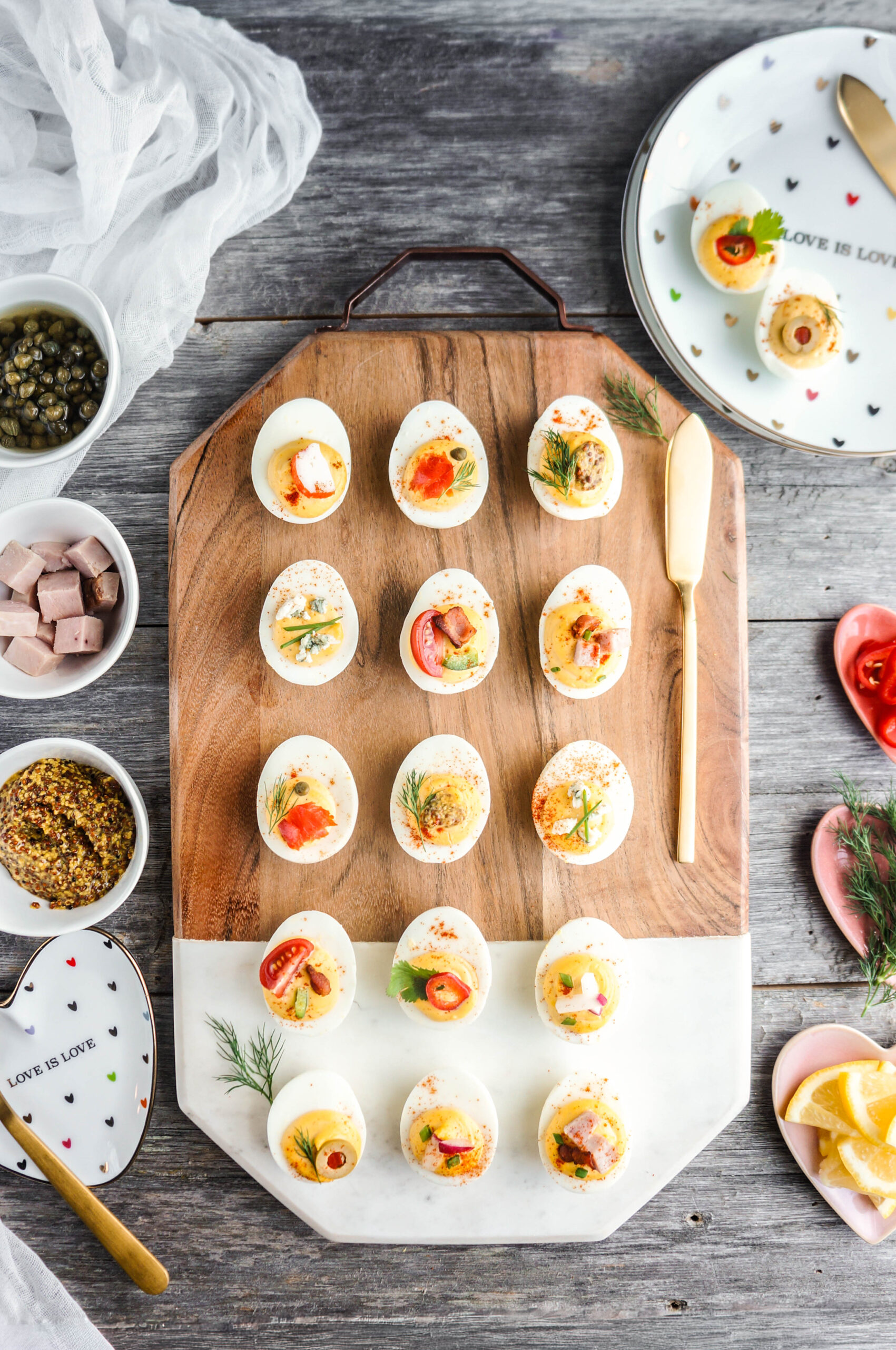 Overhead view of a deviled egg platter arranged on a wooden and marble serving board, featuring assorted toppings like smoked salmon, dill, capers, tomatoes, chili peppers, bacon, blue cheese, and olives. Small bowls of capers, diced ham, whole-grain mustard, and sliced peppers surround the board, along with decorative heart plates reading ‘LOVE IS LOVE,’ creating a bright and inviting appetizer display.