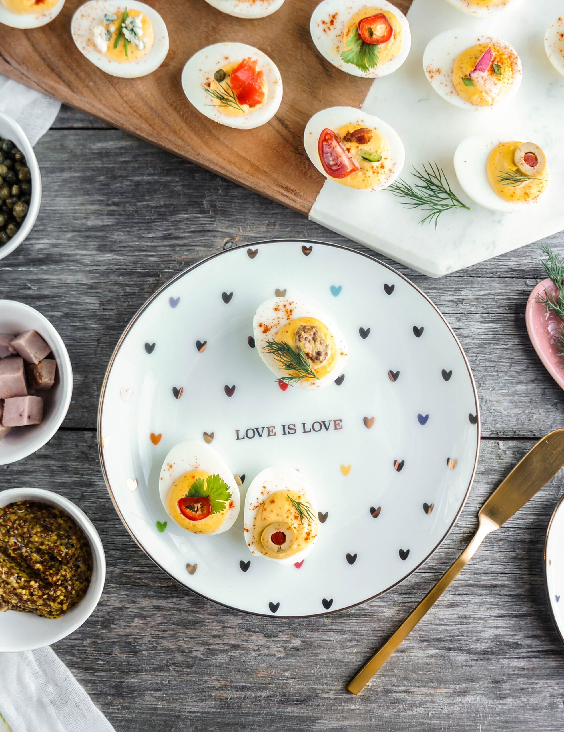 The ultimate party platter: A Deviled Egg Bar A colorful deviled egg platter displayed on a wooden and marble board, featuring assorted toppings like smoked salmon, dill, capers, blue cheese, bacon, tomatoes, chili peppers, and herbs. Three deviled eggs are plated on a white dish decorated with tiny multicolored hearts and the phrase ‘LOVE IS LOVE,’ surrounded by small bowls of diced ham, whole-grain mustard, and capers. A gold butter knife rests alongside the plate on a rustic wooden surface