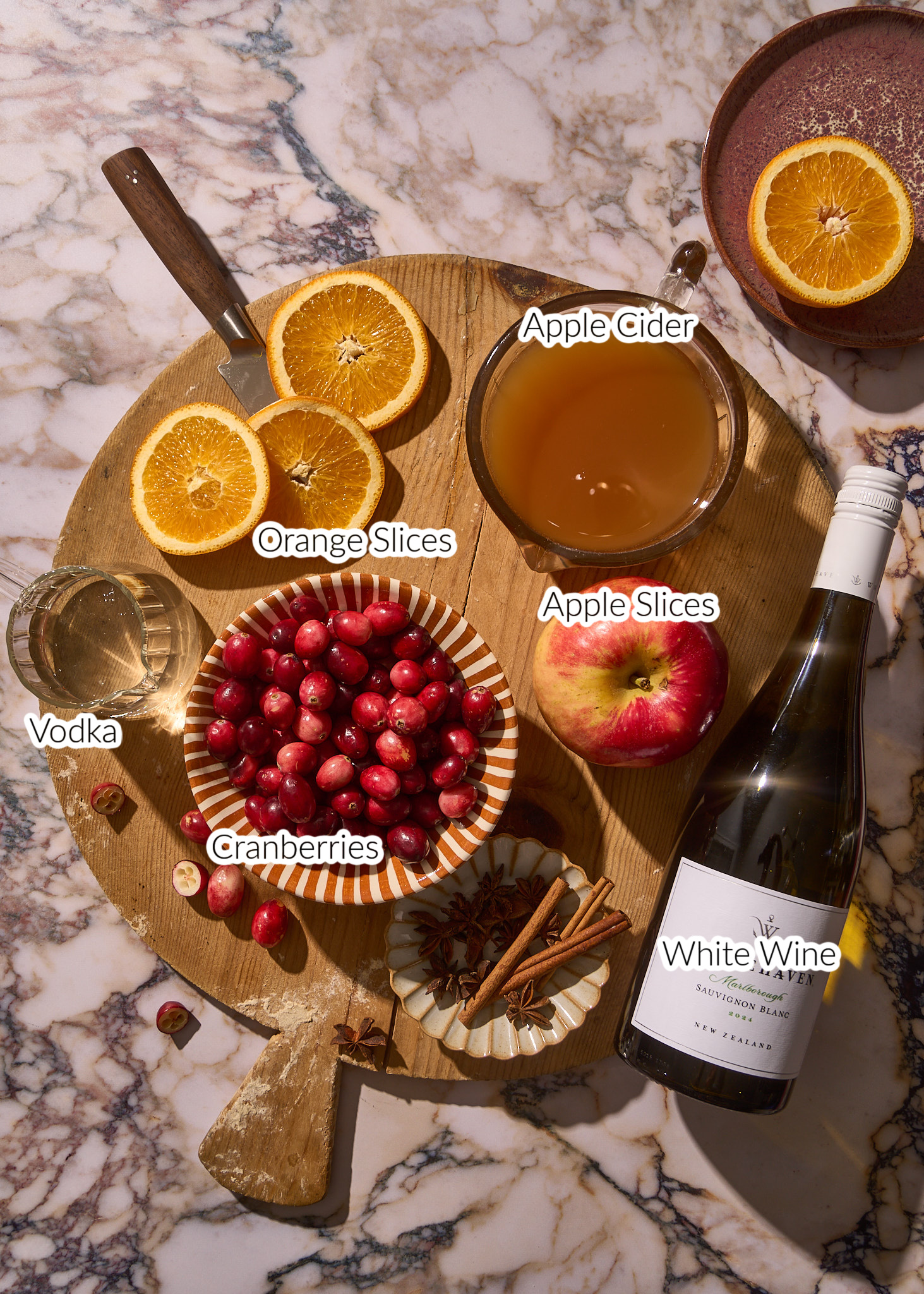 Here is alt text using the words *apple cider sangria*:
**Alt text:**
Overhead view of ingredients for apple cider sangria arranged on a round wooden board, including fresh orange slices, a bowl of cranberries, a whole apple, a glass of apple cider, a small glass of vodka, cinnamon sticks, and a bottle of Sauvignon Blanc white wine, all styled on a marble countertop with warm, cozy autumn lighting.