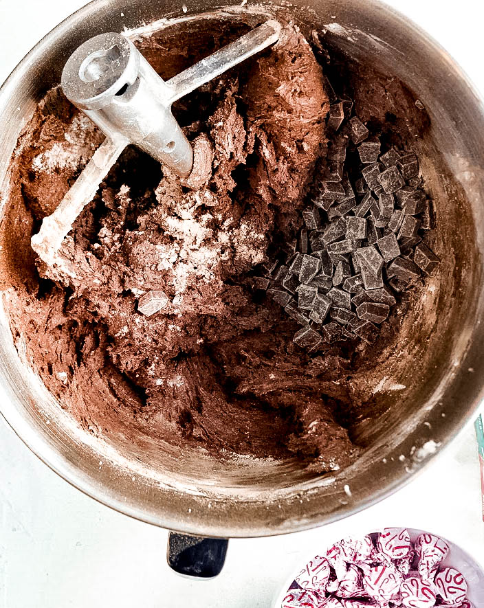 Double Chocolate Peppermint Kiss Cookies Chocolate cookie dough for Peppermint chocolate kiss cookies being mixed in a stand mixer bowl, with a pile of chocolate chunks ready to be stirred in. A small bowl of unwrapped peppermint chocolate kisses sits nearby, showing the ingredients in progress.