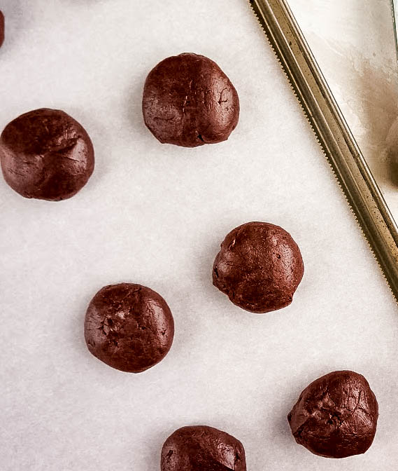 Double Chocolate Peppermint Kiss Cookies Chocolate dough balls for Peppermint chocolate kiss cookies arranged on a parchment-lined baking sheet, ready to go into the oven before being topped with peppermint chocolate kisses.