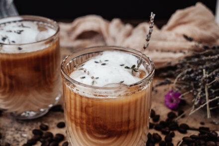 iced lavender latte on wood surface surrounded by coffee beans and dried lavender in the background