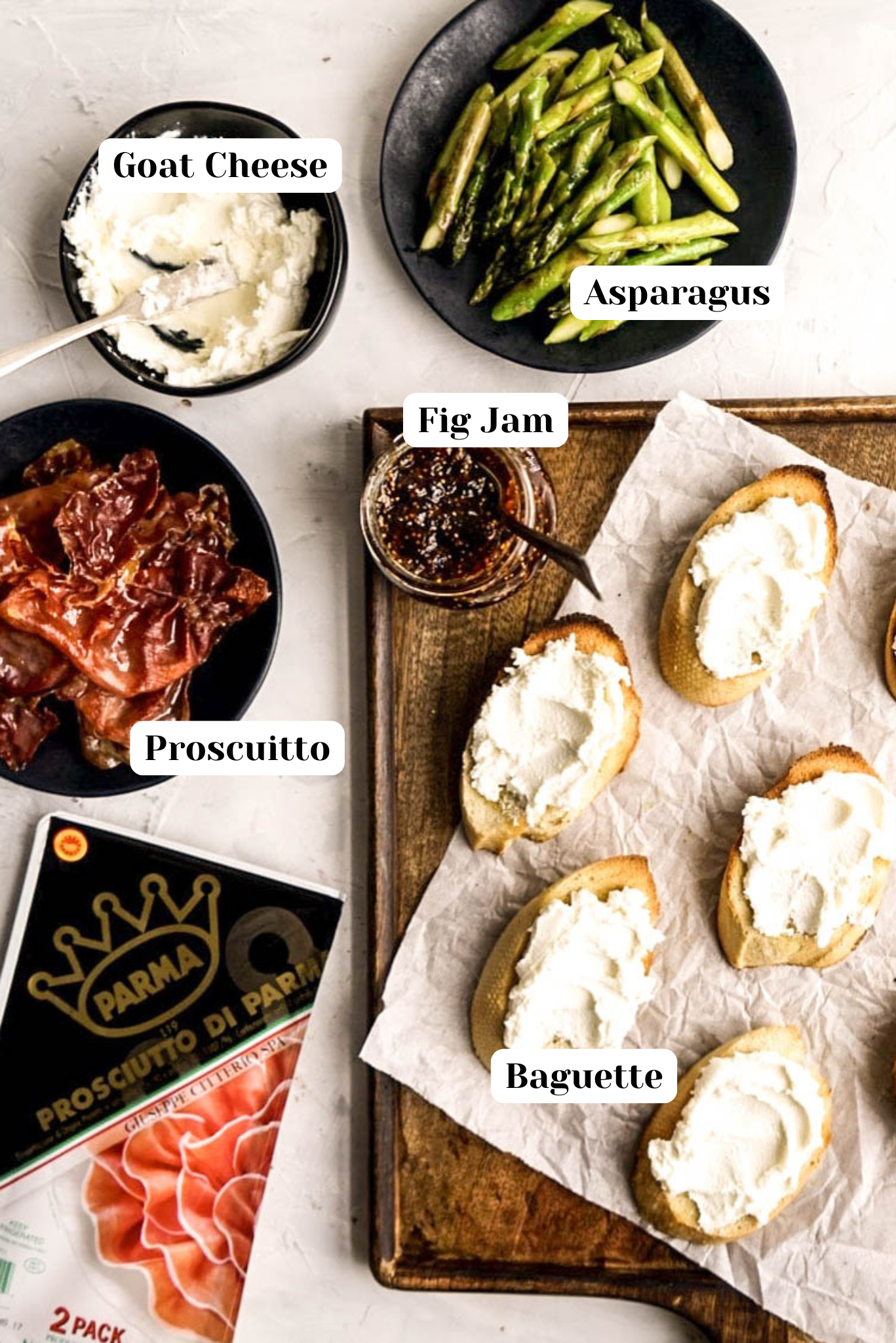 Overhead view of prosciutto crostini ingredients including goat cheese, roasted asparagus, fig jam, sliced baguette with goat cheese, and prosciutto di Parma arranged on a light surface for assembling prosciutto crostini.