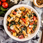 Overhead view of rigatoni with sausage and tomatoes in a white bowl, tossed with wilted spinach, juicy cherry tomatoes, and Italian sausage, finished with shaved Parmesan and red pepper flakes on a light marble surface.