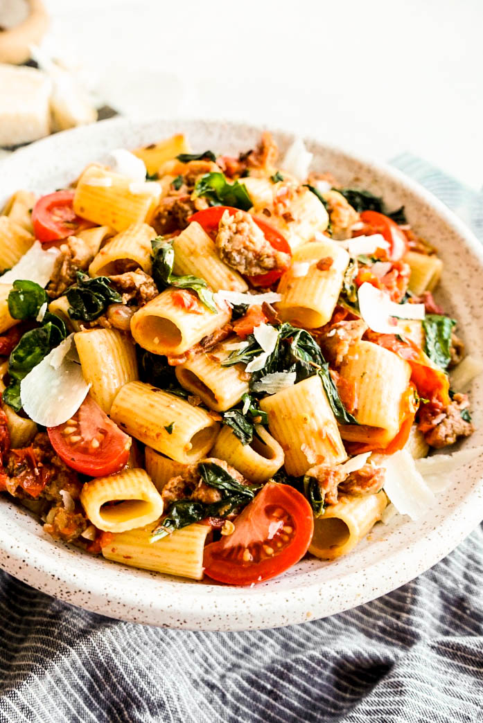 Close-up side view photo of rigatoni with sausage and tomatoes served in a shallow bowl, with Italian sausage crumbles, cherry tomatoes, fresh spinach, and Parmesan cheese.