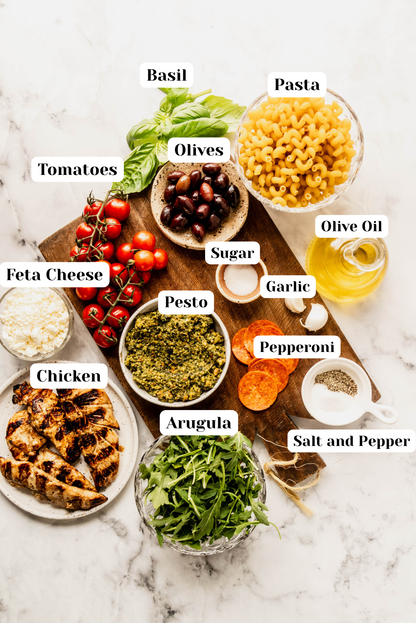 Overhead ingredient shot for pasta salad pesto chicken featuring labeled items including basil, pasta, olives, cherry tomatoes, feta cheese, grilled chicken, pesto, arugula, garlic, olive oil, pepperoni, sugar, and salt and pepper on a marble surface.