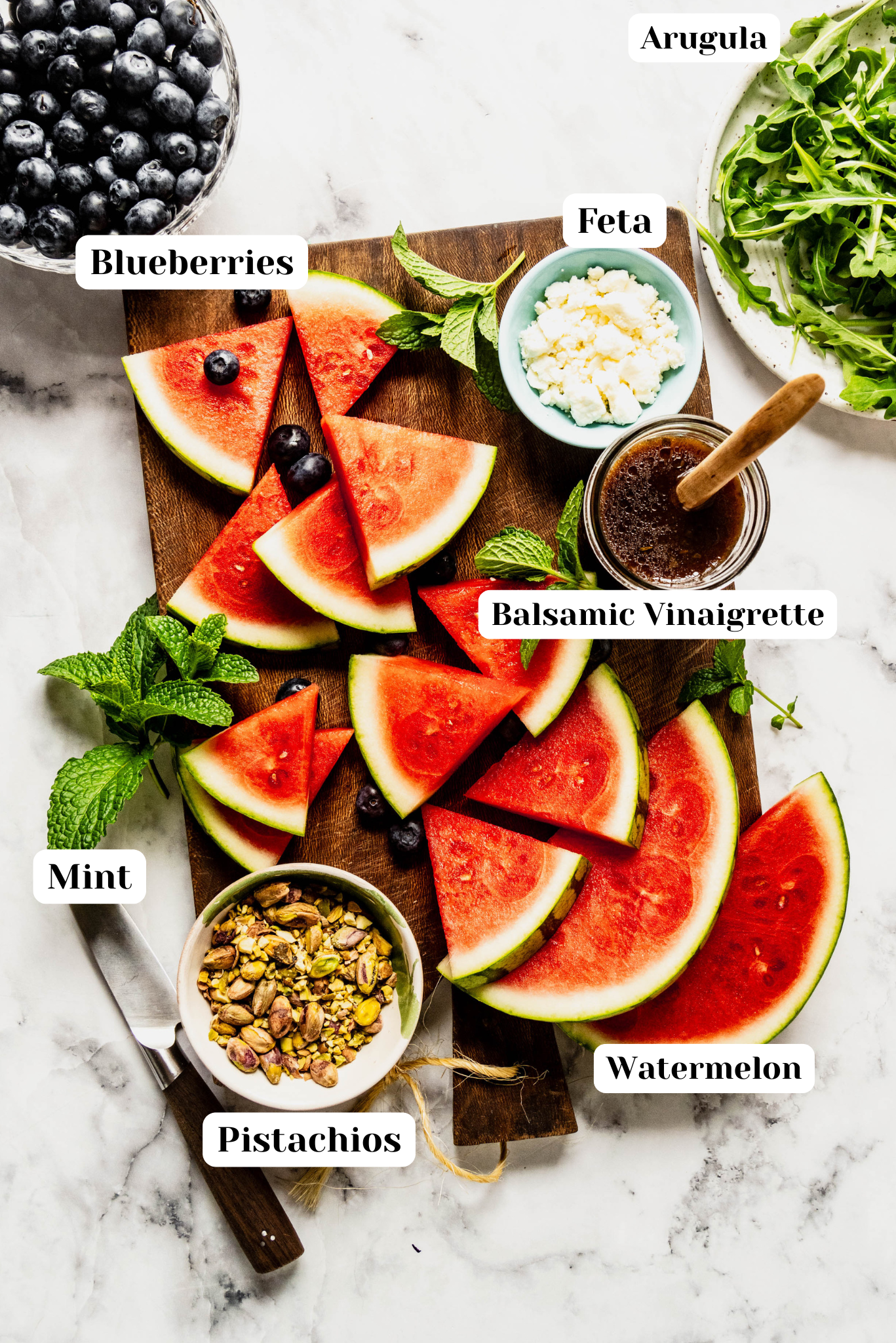 Ingredient shot for an arugula and watermelon salad featuring fresh watermelon slices, blueberries, crumbled feta, baby arugula, mint, pistachios, and balsamic vinaigrette arranged on a wooden board over a white marble surface with ingredient labels.