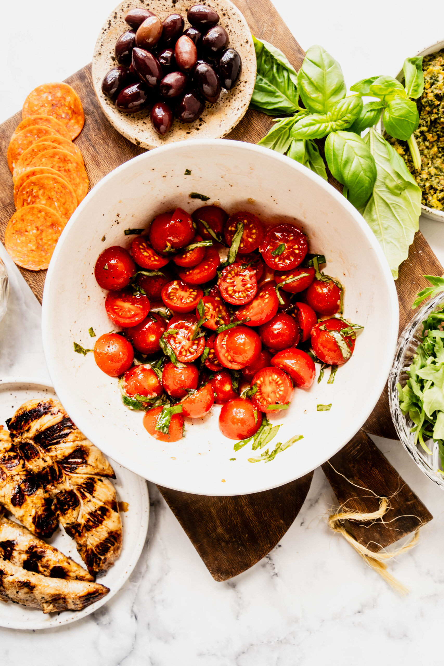 Overhead view of bruschetta tomatoes being prepared for pasta salad pesto chicken, featuring halved cherry tomatoes tossed with basil, alongside grilled chicken, olives, pepperoni, pesto, and fresh ingredients on a marble surface.