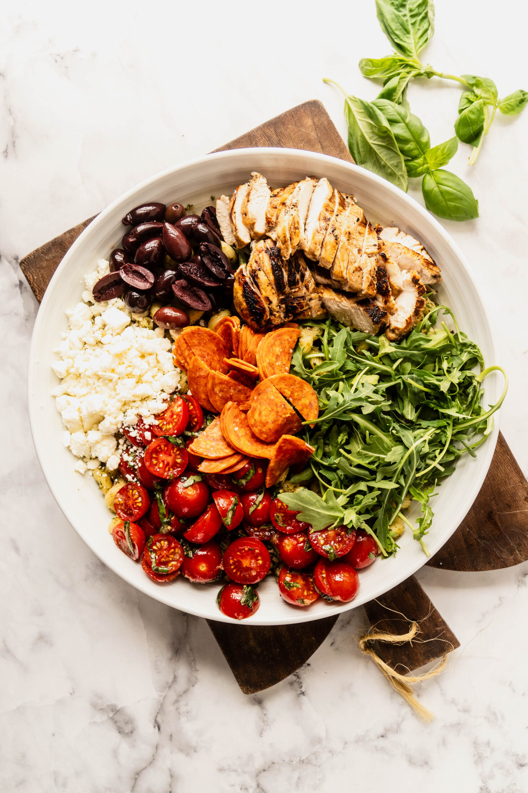 Overhead view of pasta salad pesto chicken arranged in a bowl with grilled sliced chicken, bruschetta tomatoes, pepperoni, feta cheese, kalamata olives, and fresh arugula over pesto-coated pasta on a marble background.