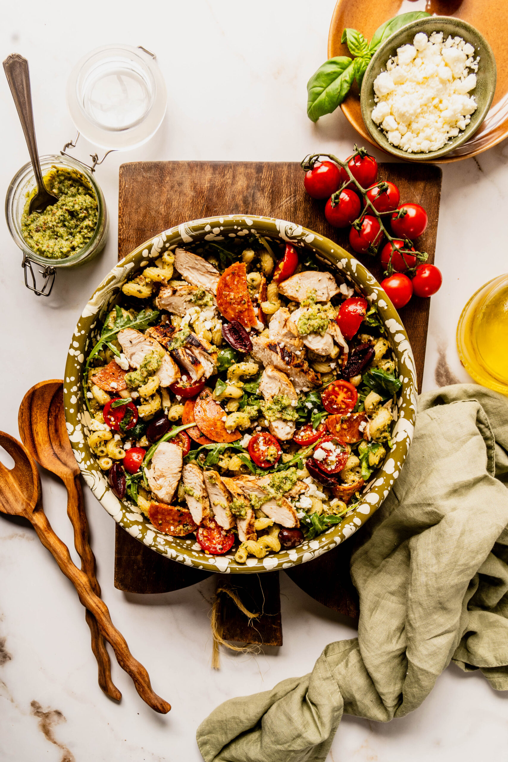 Overhead view of pasta salad pesto chicken in a large serving bowl with corkscrew pasta tossed in basil pesto, grilled chicken slices, cherry tomatoes, kalamata olives, pepperoni, feta cheese, and arugula, on a marble surface.