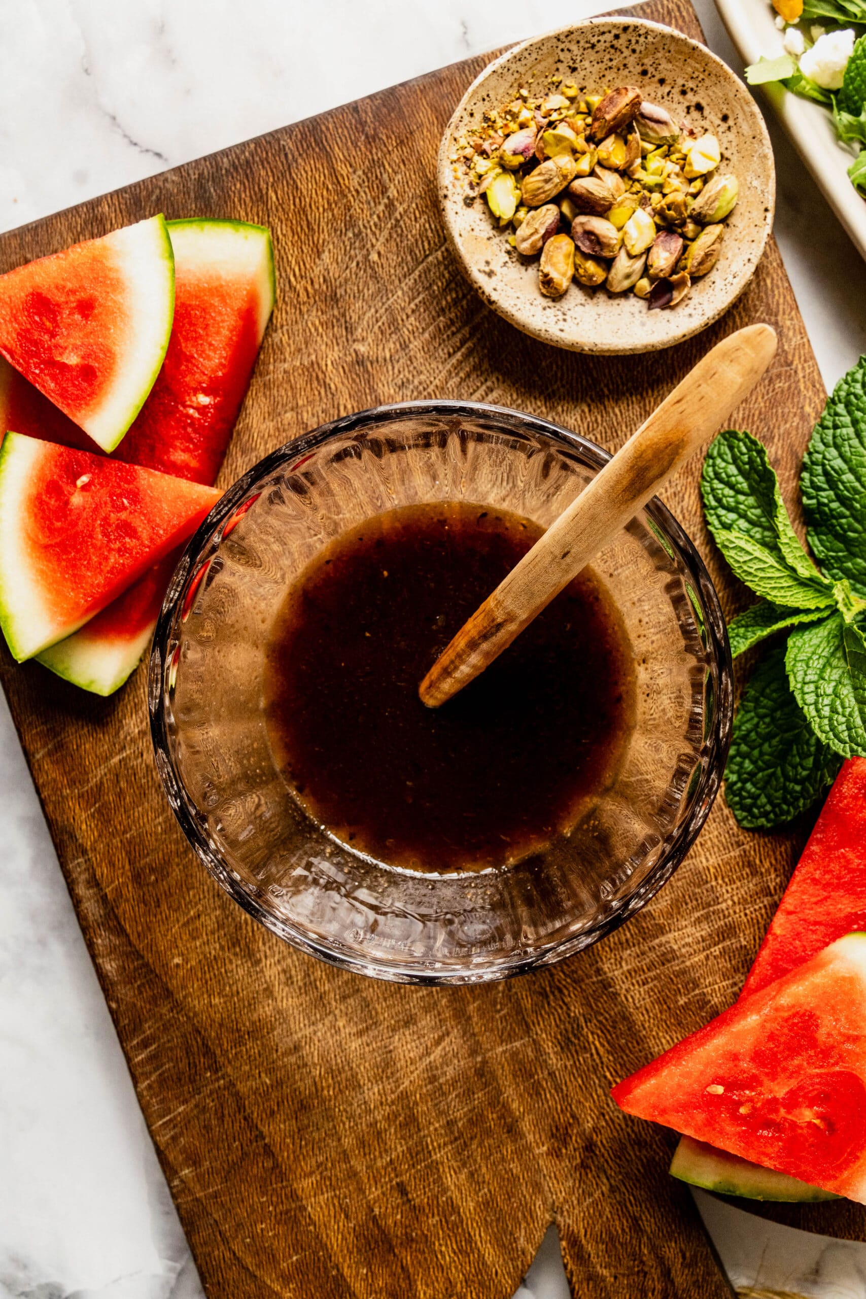 Close-up of homemade balsamic vinaigrette for an arugula and watermelon salad, served in a glass bowl with fresh watermelon wedges, pistachios, and mint on a rustic wooden board over a white marble surface.