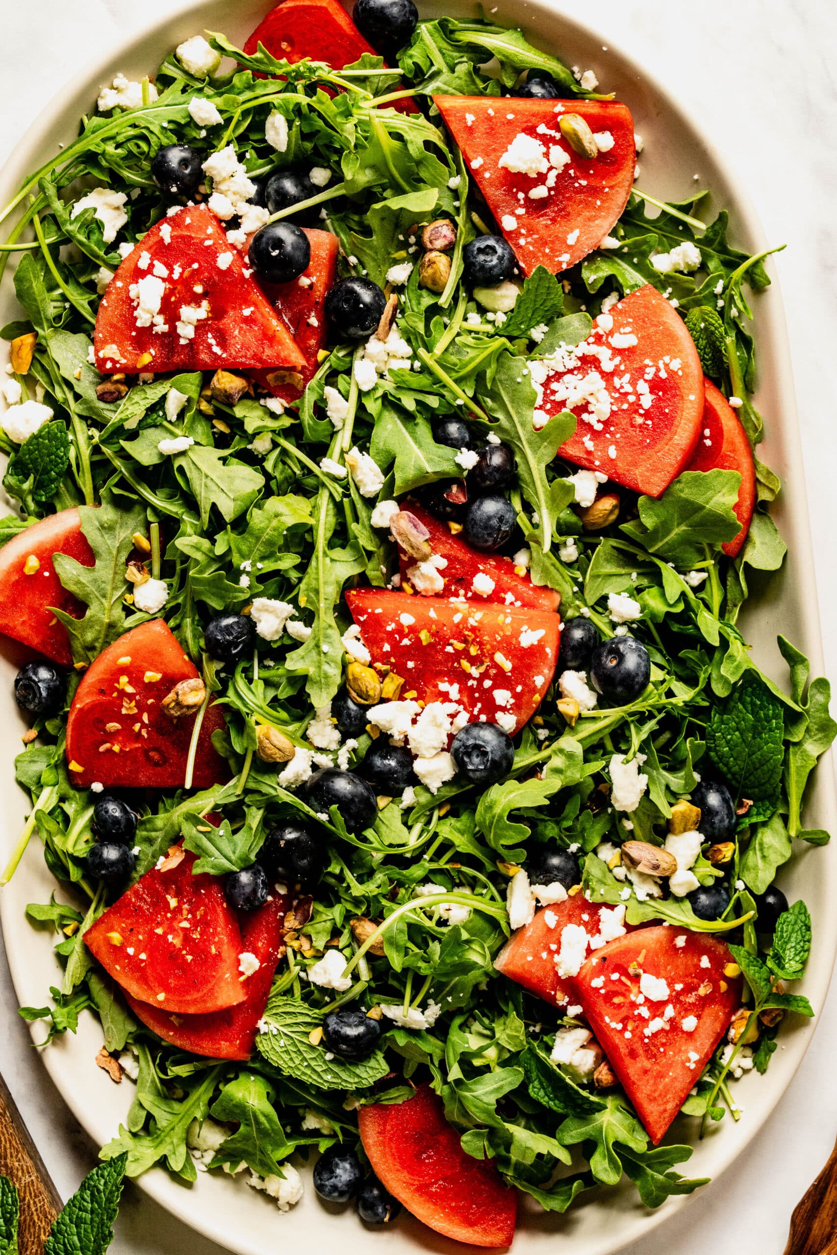 Close-up overhead view of an arugula and watermelon salad made with fresh arugula, juicy watermelon wedges, blueberries, crumbled feta cheese, chopped pistachios, and mint leaves on a large serving platter, creating a colorful and refreshing summer salad.