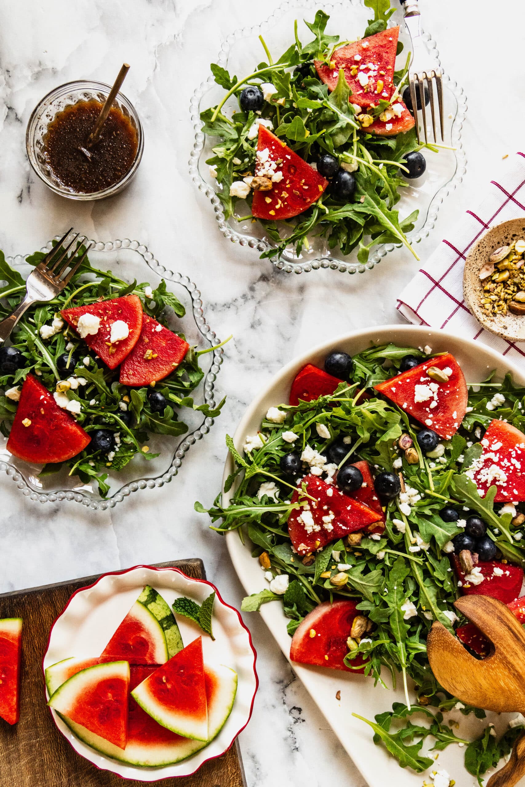 Overhead view of an arugula and watermelon salad served family-style on a large platter with individual salad plates, featuring fresh arugula, juicy watermelon wedges, blueberries, crumbled feta cheese, pistachios, and mint, alongside balsamic vinaigrette on a white marble table.