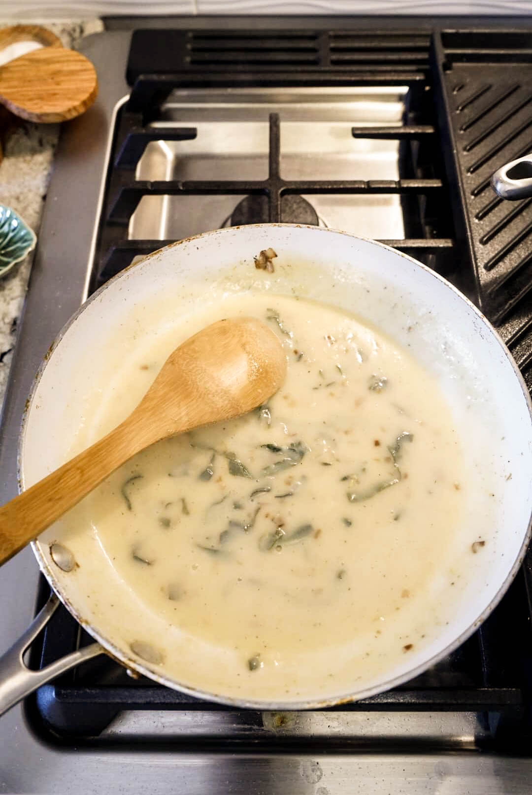 Creamy Milanese sauce with chopped sage simmering in a skillet on the stovetop, being stirred with a wooden spoon.