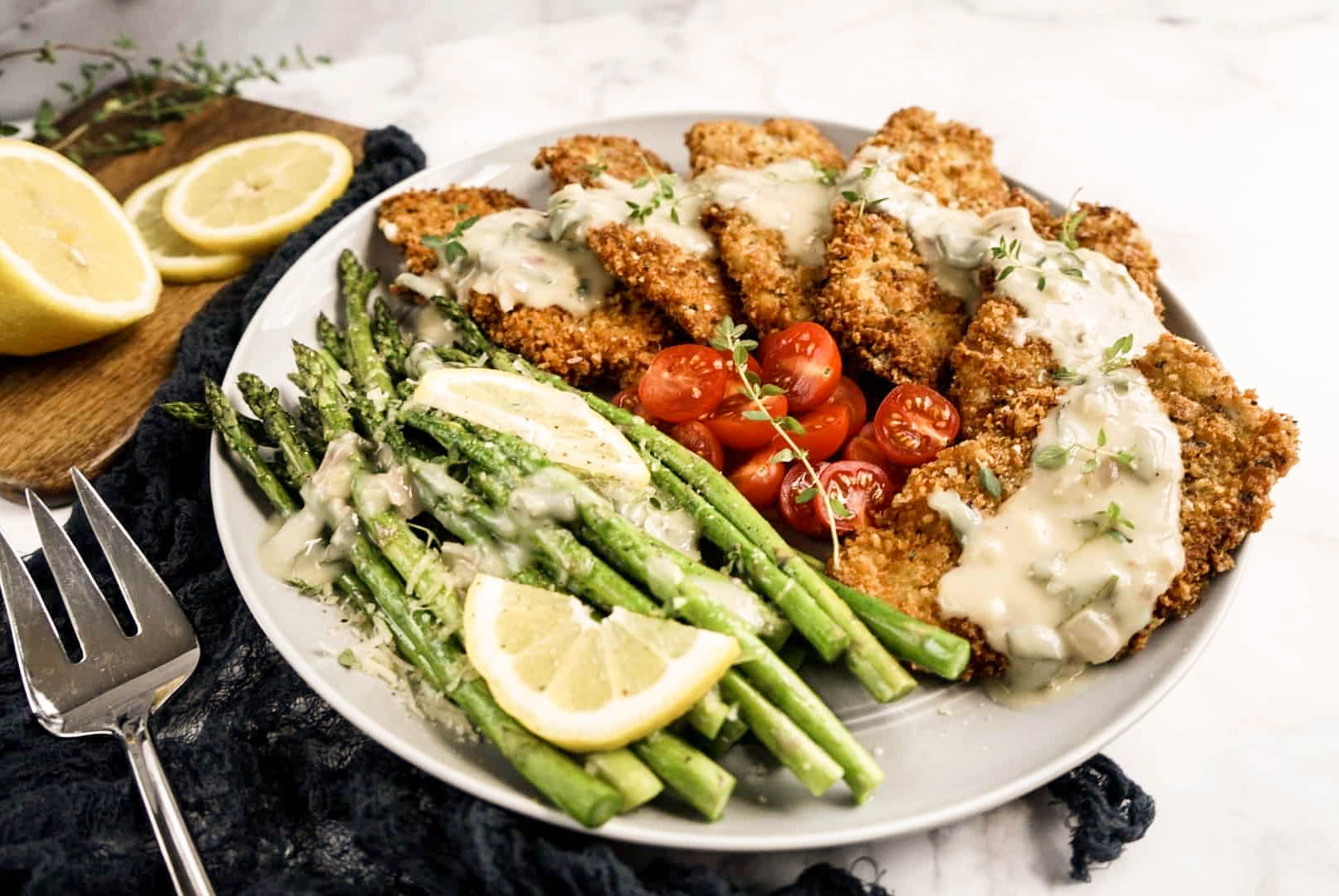 Crispy breaded veal Milanese served on a platter with roasted asparagus, cherry tomatoes, and lemon slices, topped with creamy Milanese sauce and fresh herbs.