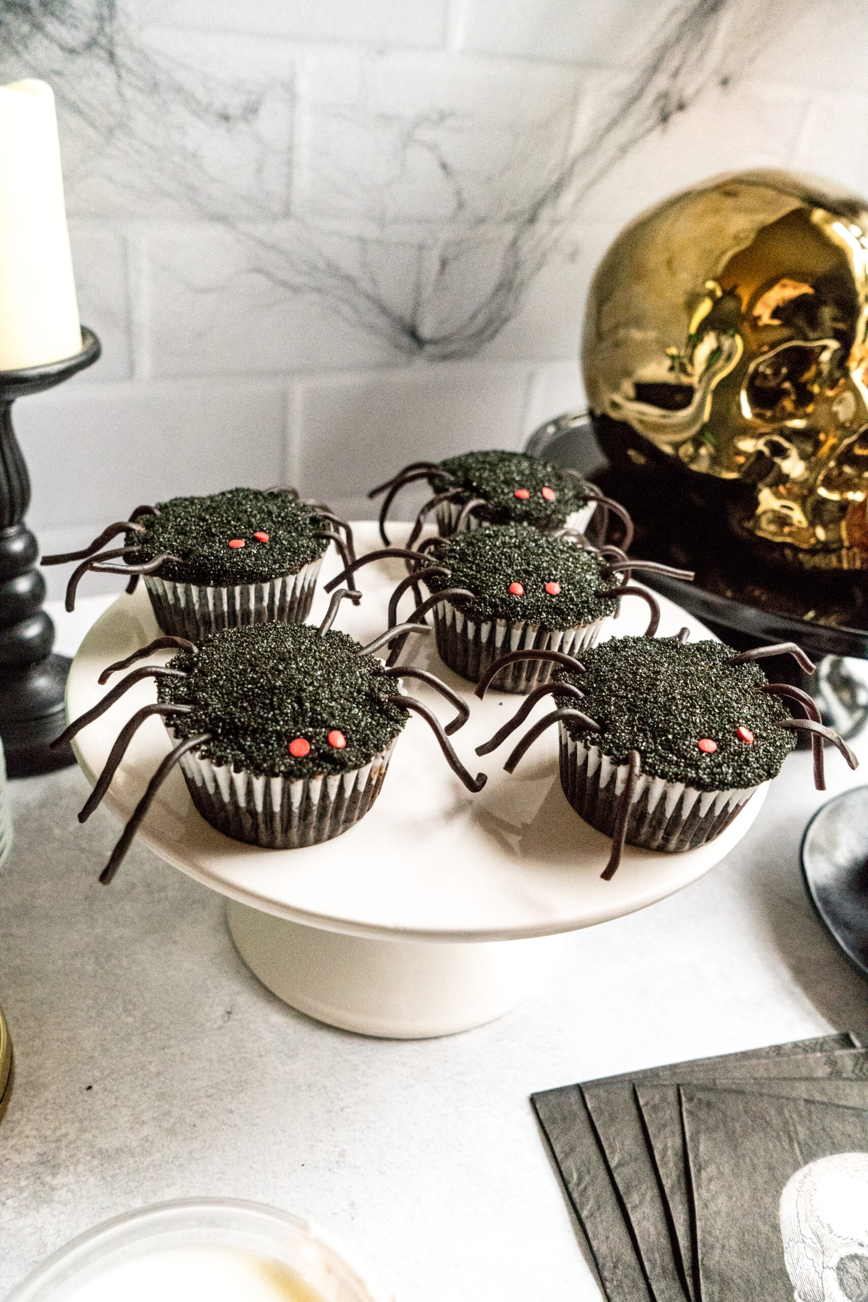 Close-up of Halloween spider cupcakes on a white cake stand. Each cupcake is covered in black sprinkles with red candy eyes and black licorice legs. The spooky setup includes cobweb decorations, a gold skull, and black napkins, creating a festive Halloween atmosphere.