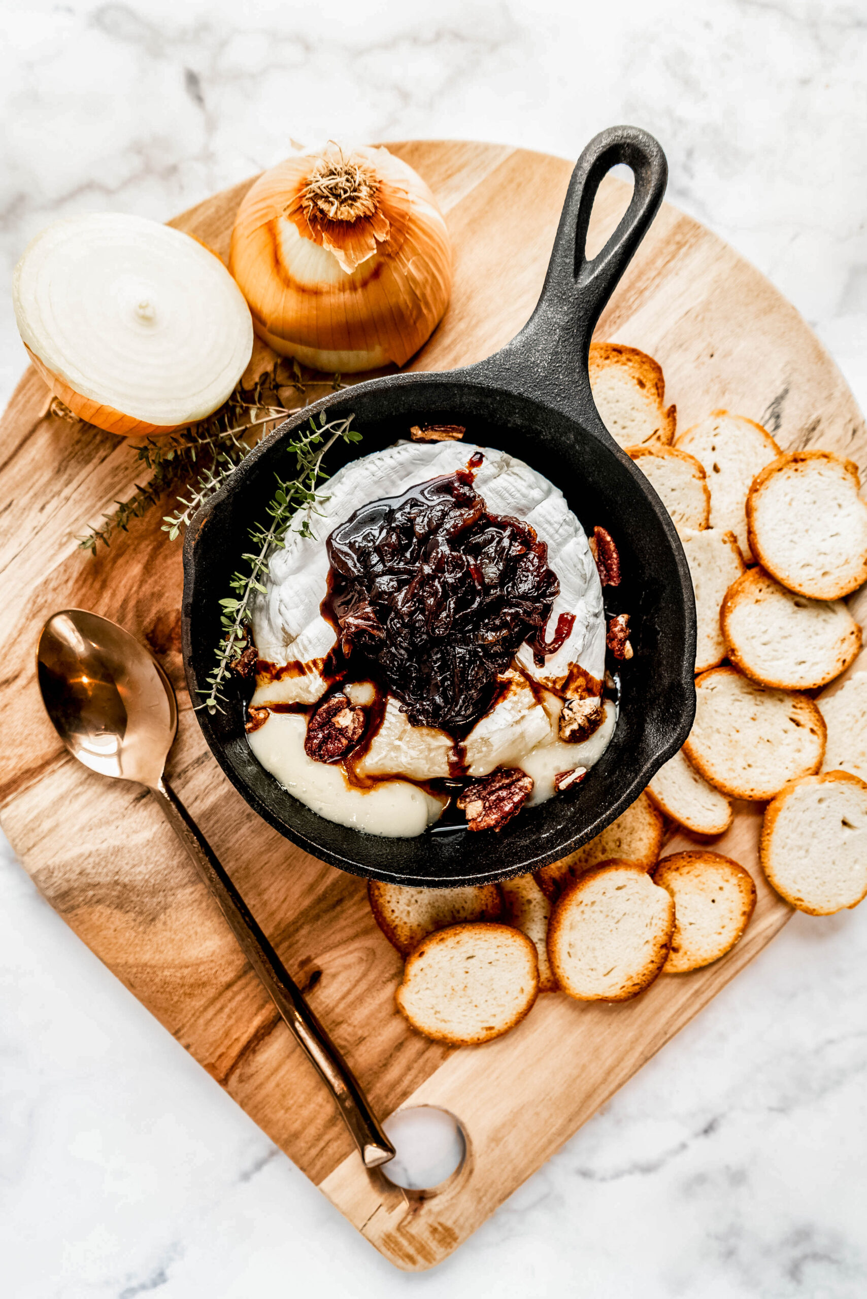 Overhead view of baked brie in a small cast-iron pan, topped with rich caramelized onions and pecans, served alongside toasted baguette slices on a wooden board with fresh thyme and a serving spoon.