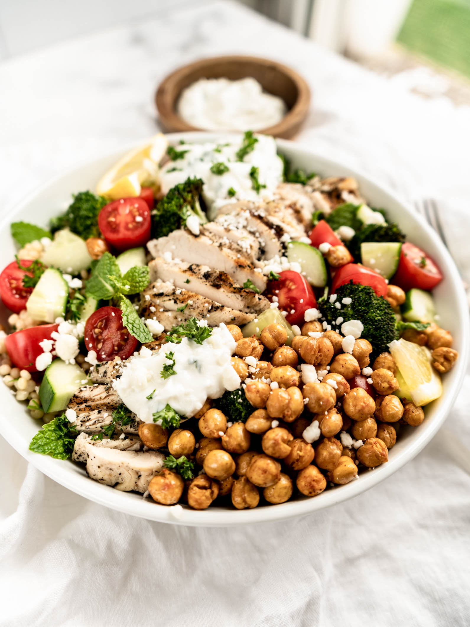 greek chicken bowl Alt text: Bright and colorful Greek chicken bowls served in a white bowl, featuring sliced grilled chicken breast, roasted chickpeas, cherry tomatoes, cucumber, broccoli, fresh herbs, crumbled feta, and dollops of creamy tzatziki on a light marble surface.