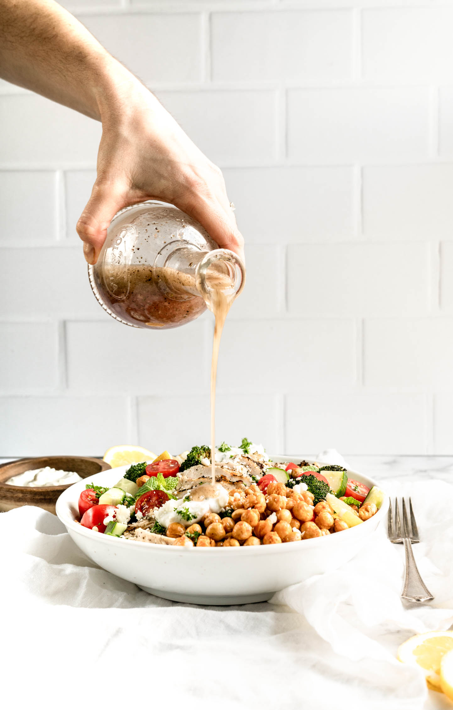 Hand pouring homemade vinaigrette over a vibrant Greek chicken bowls dish with sliced grilled chicken, roasted chickpeas, cherry tomatoes, cucumber, broccoli, fresh herbs, feta cheese, and creamy tzatziki in a white bowl on a marble surface.