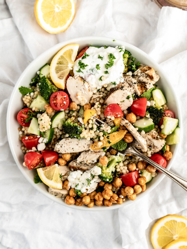 greek chicken bowl Overhead view of colorful Greek chicken bowls filled with sliced grilled chicken, pearl couscous, roasted chickpeas, cherry tomatoes, cucumber, broccoli, feta cheese, fresh herbs, lemon wedges, and a generous scoop of creamy tzatziki in a white bowl on a light marble surface.