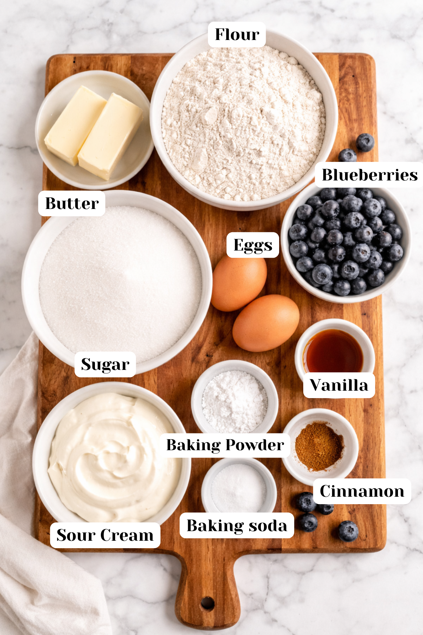 Overhead view of ingredients for sour cream blueberry muffins arranged in individual bowls on a wooden cutting board over a white marble background, including butter, sugar, eggs, sour cream, flour, blueberries, vanilla, cinnamon, baking powder, and salt.
