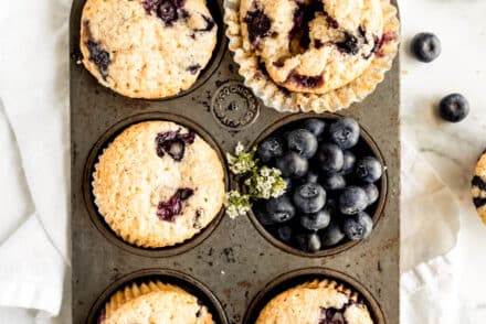 Overhead view of bakery-style blueberry sour cream muffins baked in a vintage muffin tin, with golden domed tops bursting with juicy blueberries, surrounded by fresh blueberries on a white marble background.