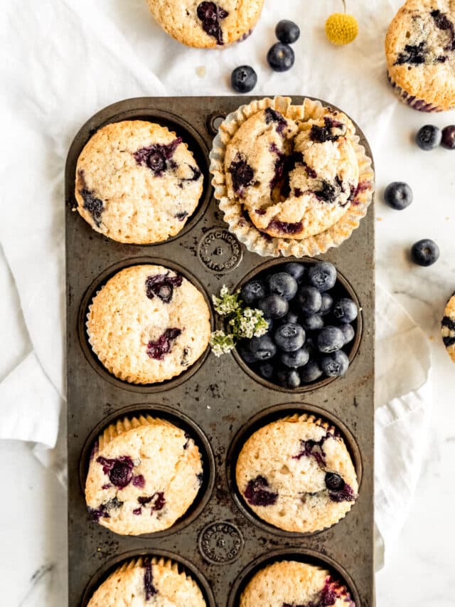 Overhead view of bakery-style blueberry sour cream muffins baked in a vintage muffin tin, with golden domed tops bursting with juicy blueberries, surrounded by fresh blueberries on a white marble background.