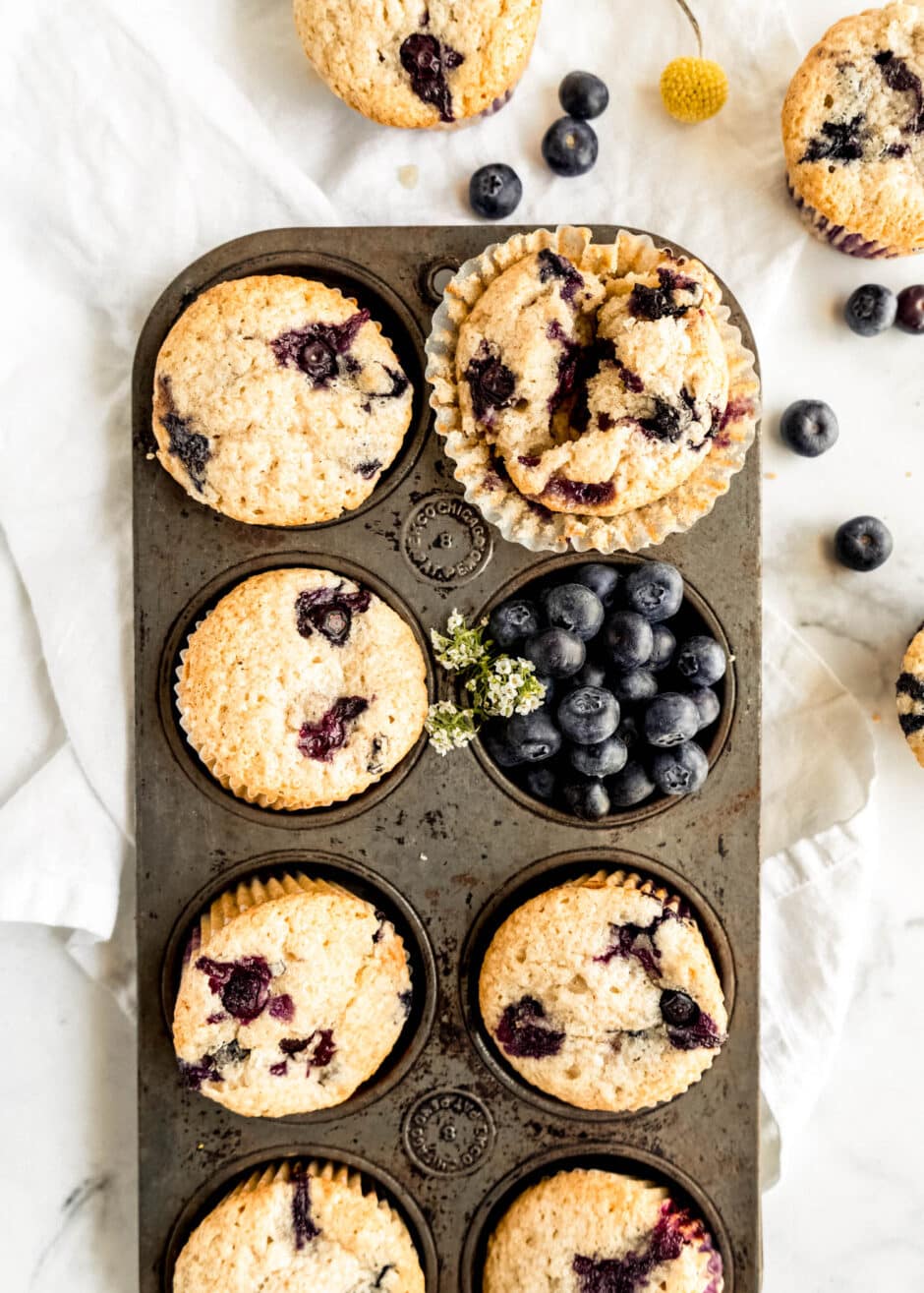 Overhead view of bakery-style blueberry sour cream muffins baked in a vintage muffin tin, with golden domed tops bursting with juicy blueberries, surrounded by fresh blueberries on a white marble background.