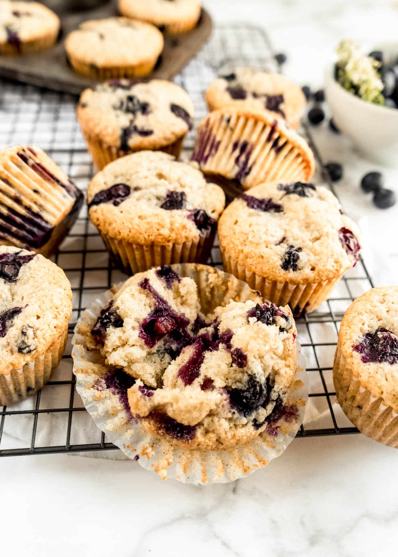Close-up of bakery-style blueberry sour cream muffins on a cooling rack, with one muffin split open to reveal a soft, tender crumb bursting with juicy blueberries, set on a white marble surface.