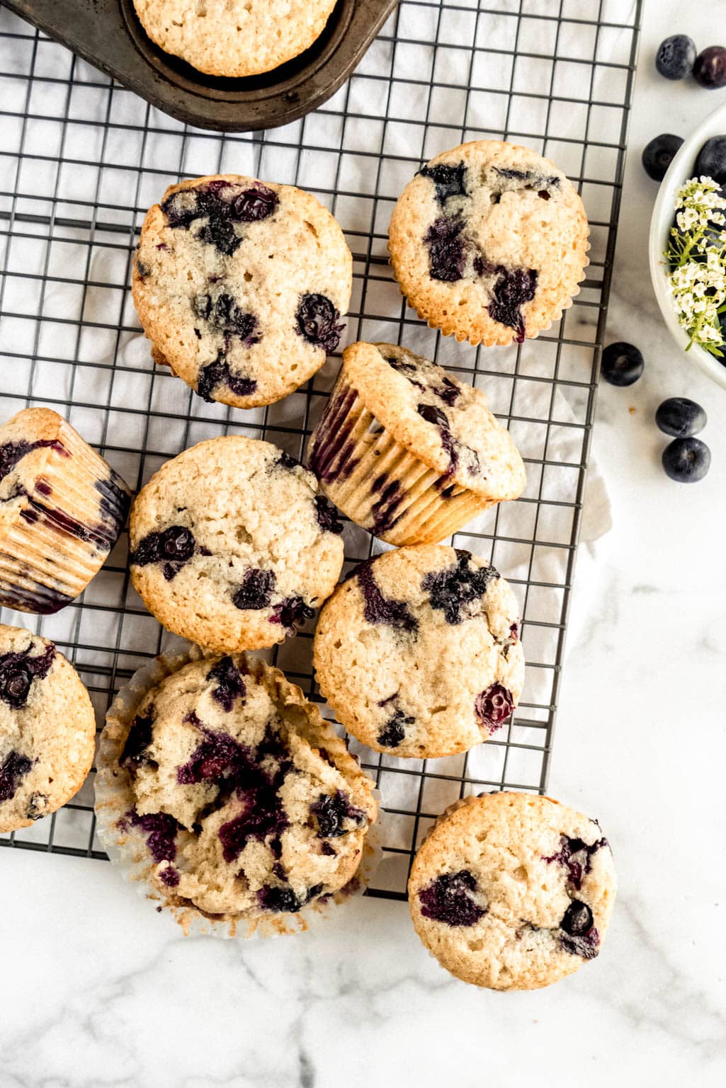 Overhead view of sour cream blueberry muffins cooling on a wire rack, featuring golden bakery-style tops with juicy blueberries and one muffin broken open to show a soft, tender crumb on a white marble background.