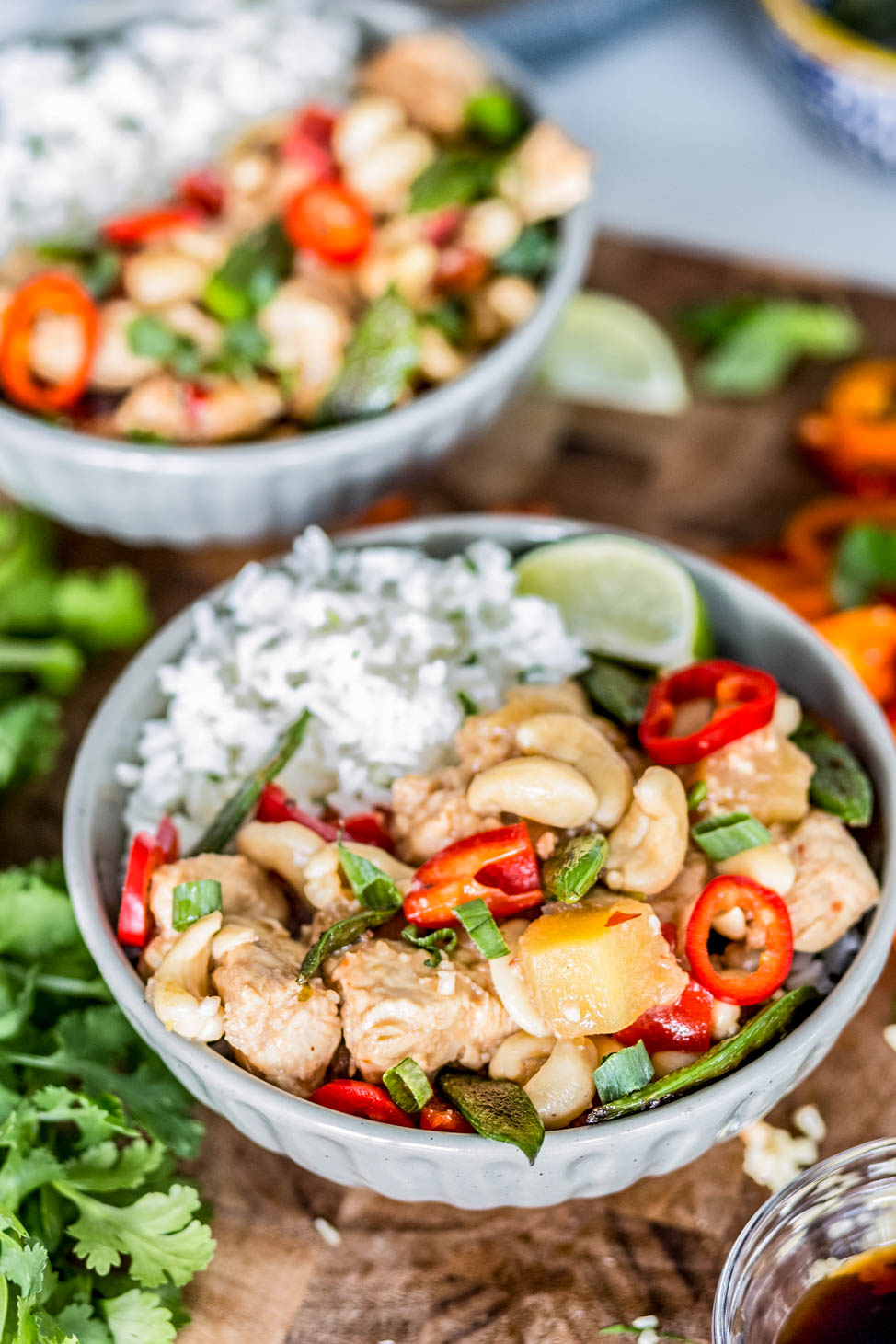 Close-up of a bowl of healthy cashew chicken with white rice, tender chicken pieces, cashews, red peppers, sugar snap peas, pineapple, and green onions, garnished with fresh herbs.