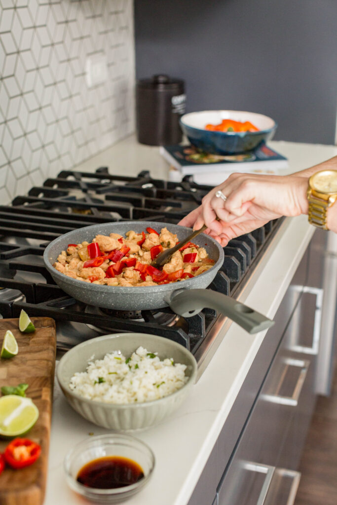 Skillet of healthy cashew chicken cooking on the stovetop with tender chicken, red bell peppers, and sauce, served alongside a bowl of white rice on a modern kitchen counter.