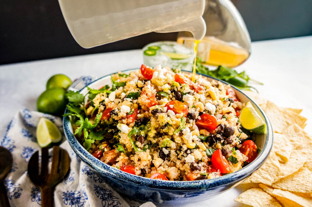 Dressing being poured over Mexican quinoa salad with black beans, cherry tomatoes, cotija cheese, fresh cilantro, and avocado, served in a bowl with tortilla chips, lime wedges, and creamy green dressing on the side
