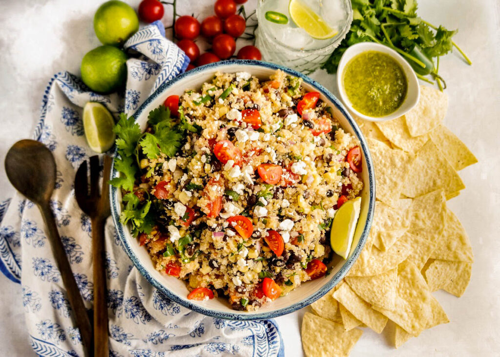 Mexican quinoa salad with black beans, cherry tomatoes, cotija cheese, fresh cilantro, and avocado, served in a bowl with tortilla chips, lime wedges, and creamy green dressing on the side
