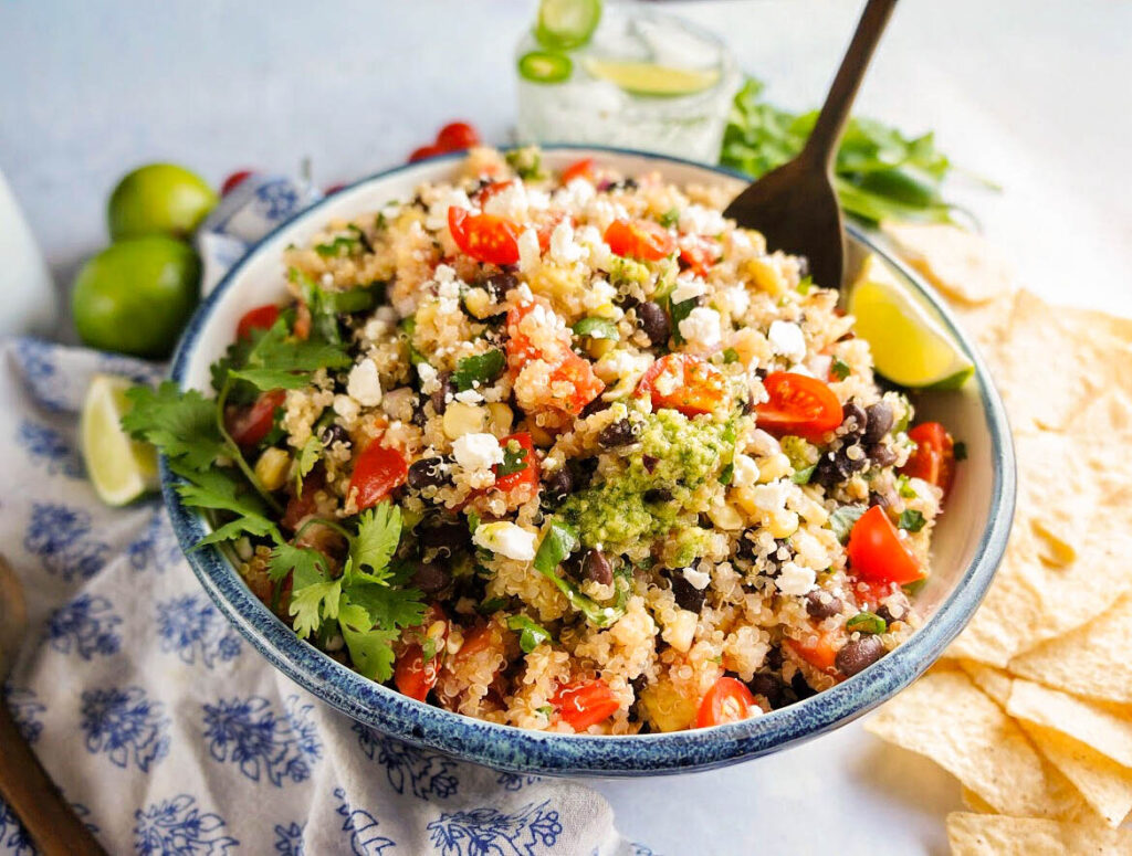 a wooden spoon placed in Mexican quinoa salad with black beans, cherry tomatoes, cotija cheese, fresh cilantro, and avocado, served in a bowl with tortilla chips, lime wedges, and creamy green dressing on the side
