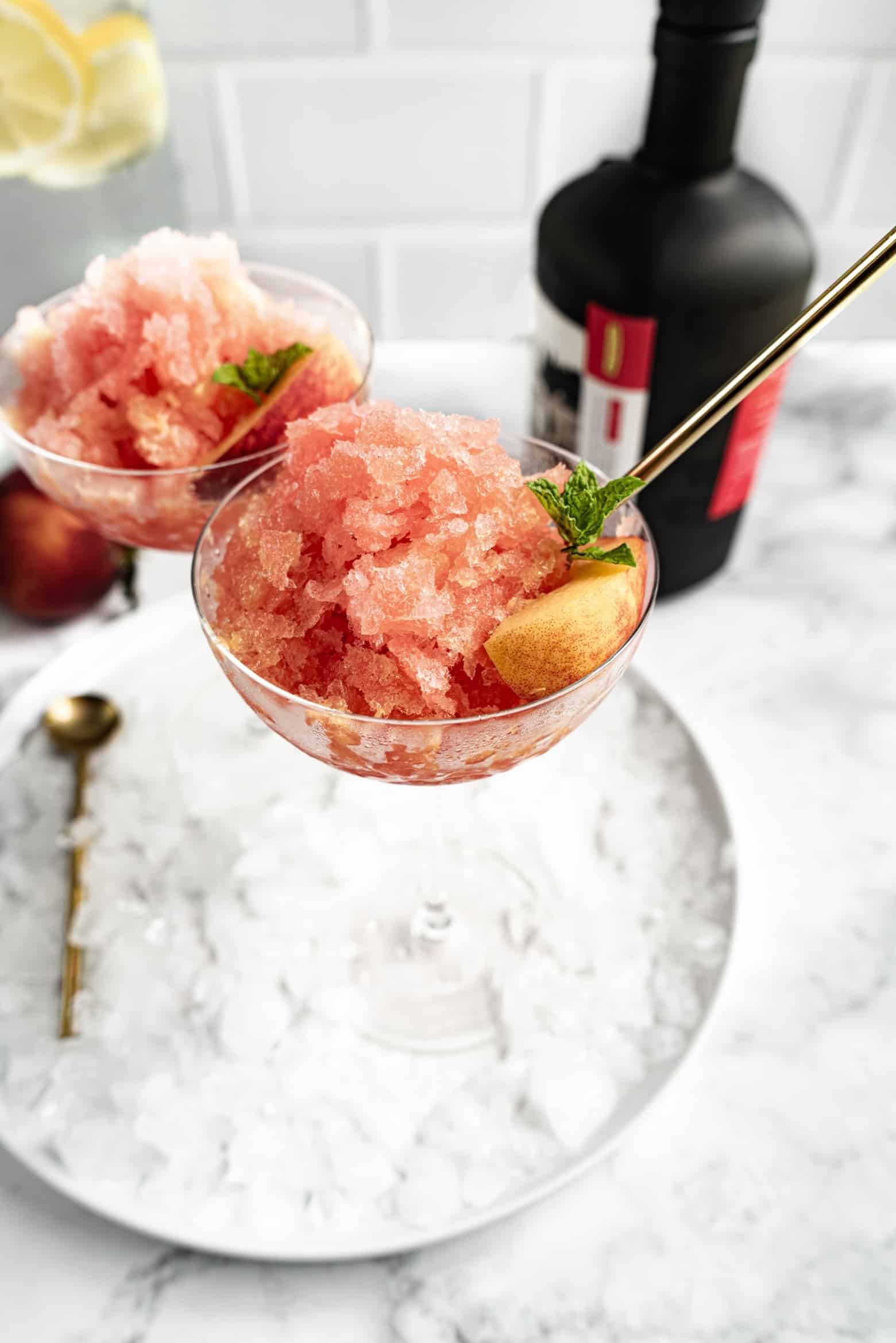 Close-up of a glass filled with peach slush, garnished with a peach slice and mint, served with a gold spoon on a tray of ice with a bottle of bourbon in the background.