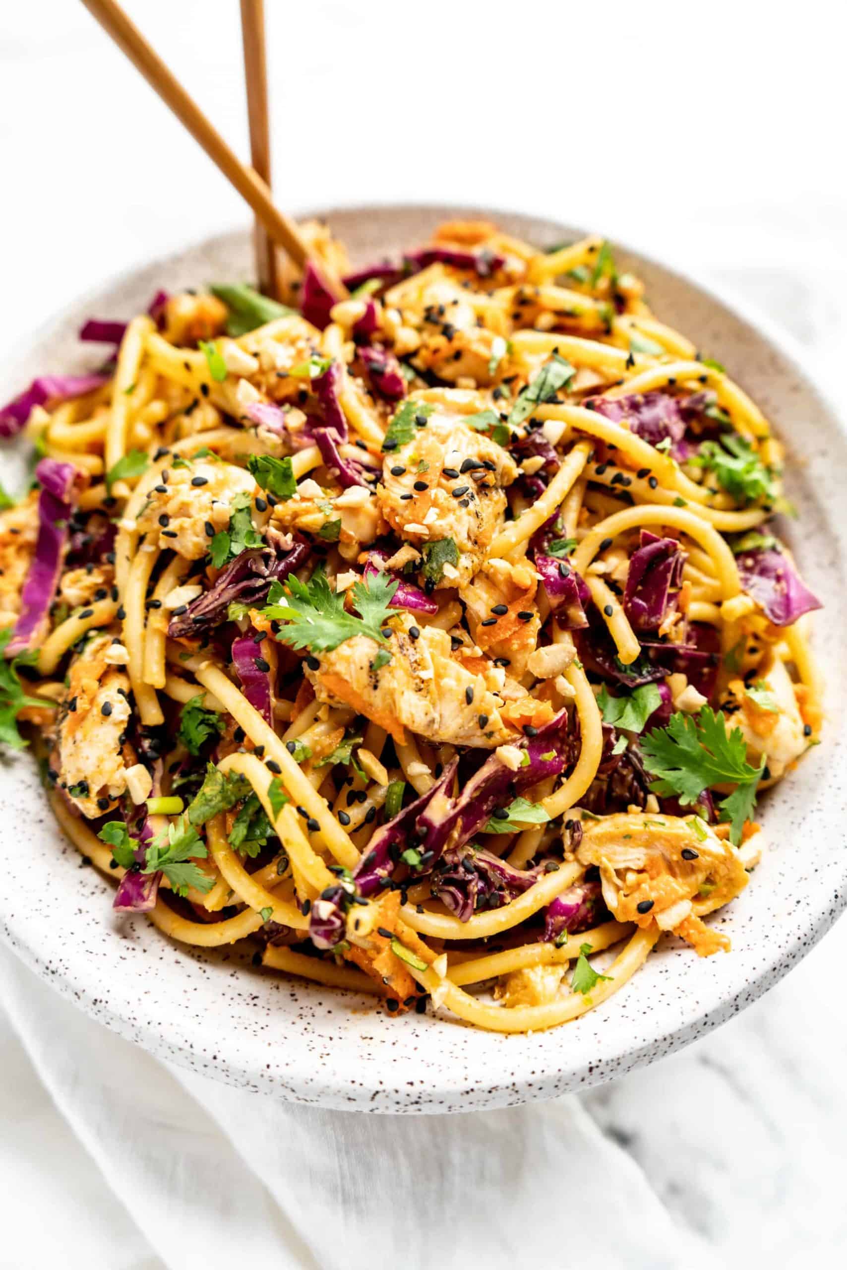 45 degree angle view of a bowl of Peanut Thai noodle salad tossed with spaghetti noodles, shredded red cabbage, carrots, chopped cilantro, tender chicken, and black sesame seeds, served with chopsticks on a white marble surface.