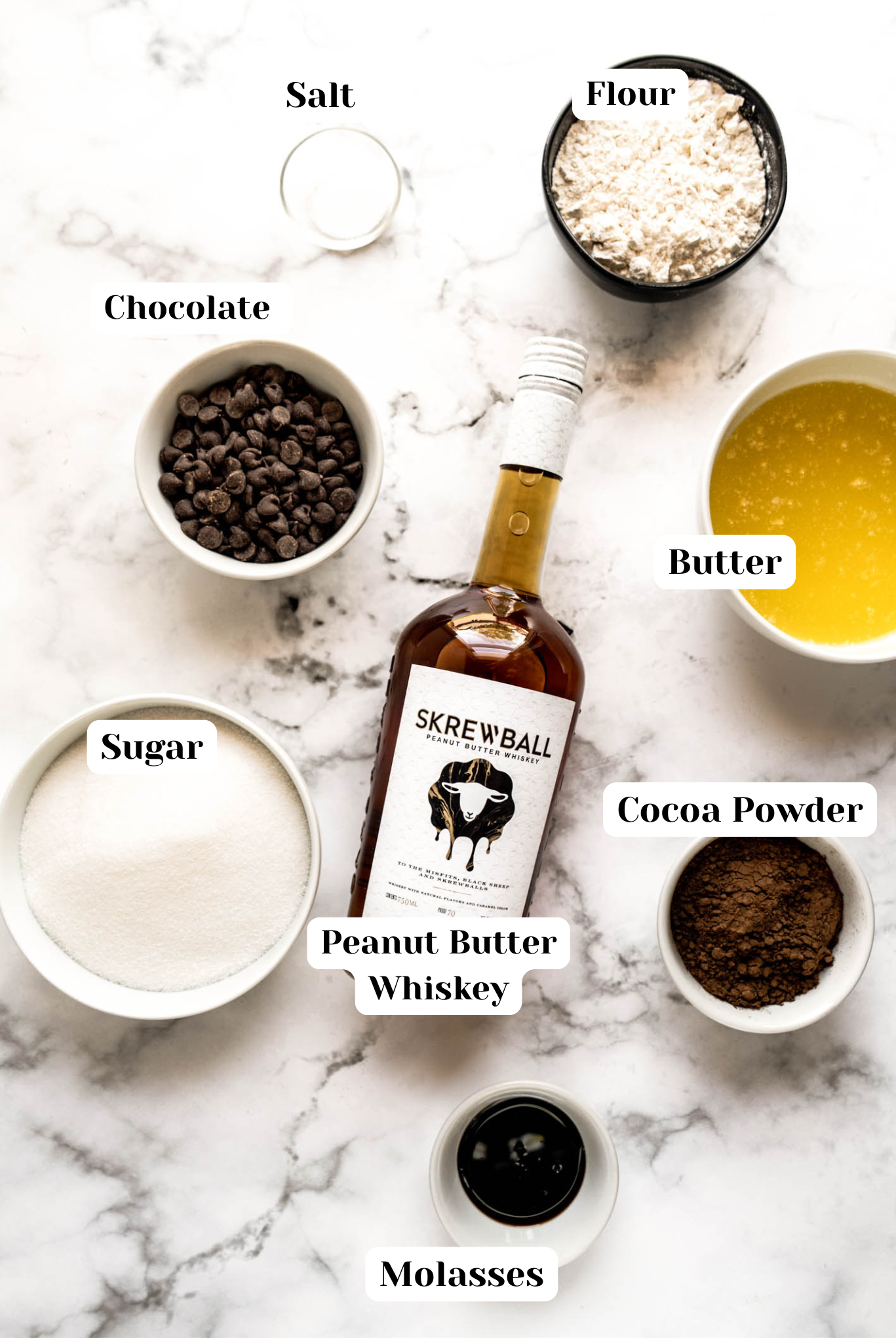 Overhead ingredient layout for peanut butter whiskey brownies including chocolate chips, sugar, flour, cocoa powder, butter, molasses, salt, and a bottle of peanut butter whiskey on a marble background.