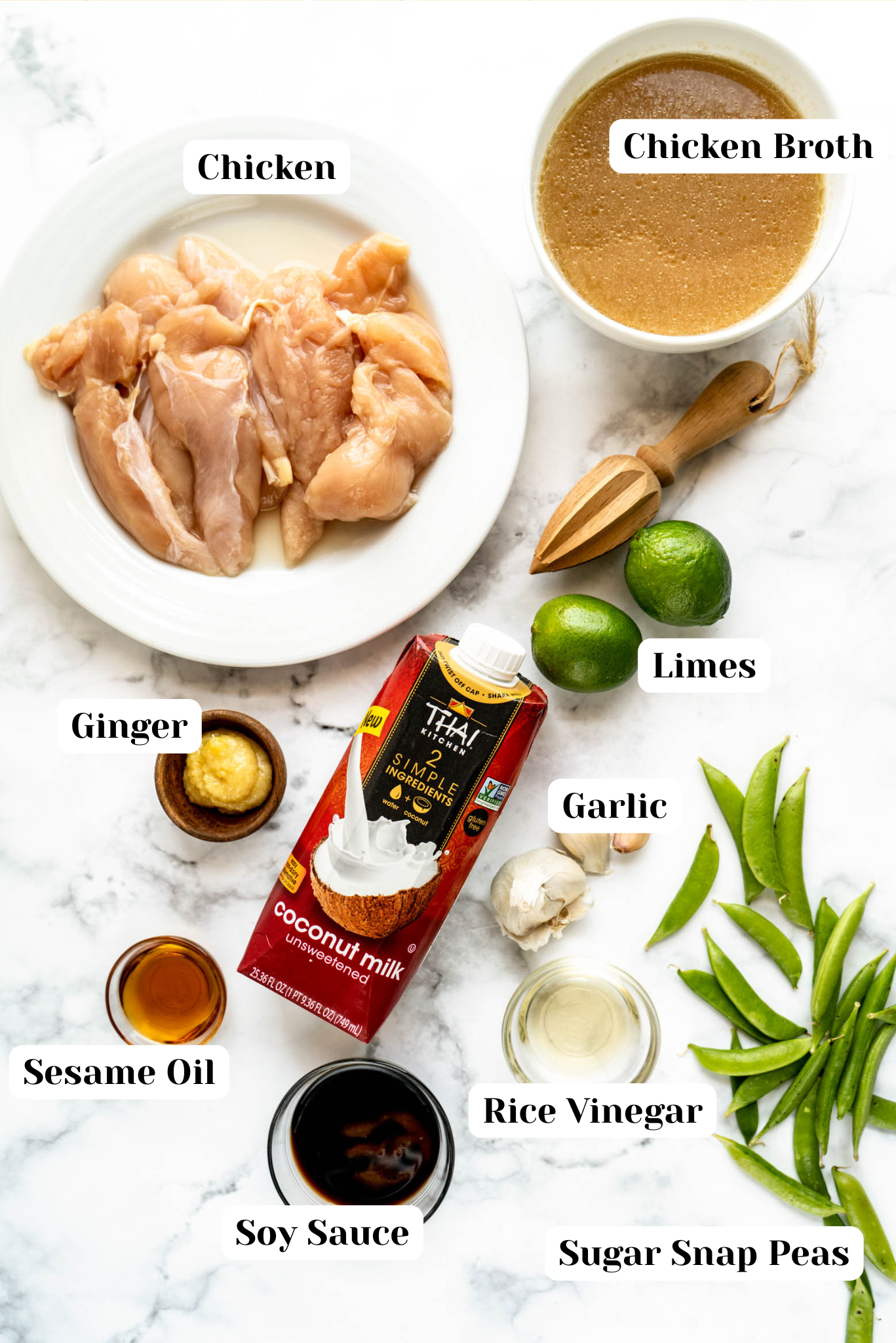 Ingredients for one pan coconut lime chicken arranged overhead on a white marble surface, including raw chicken, coconut milk, chicken broth, fresh limes, garlic, ginger, sesame oil, soy sauce, rice vinegar, and sugar snap peas.