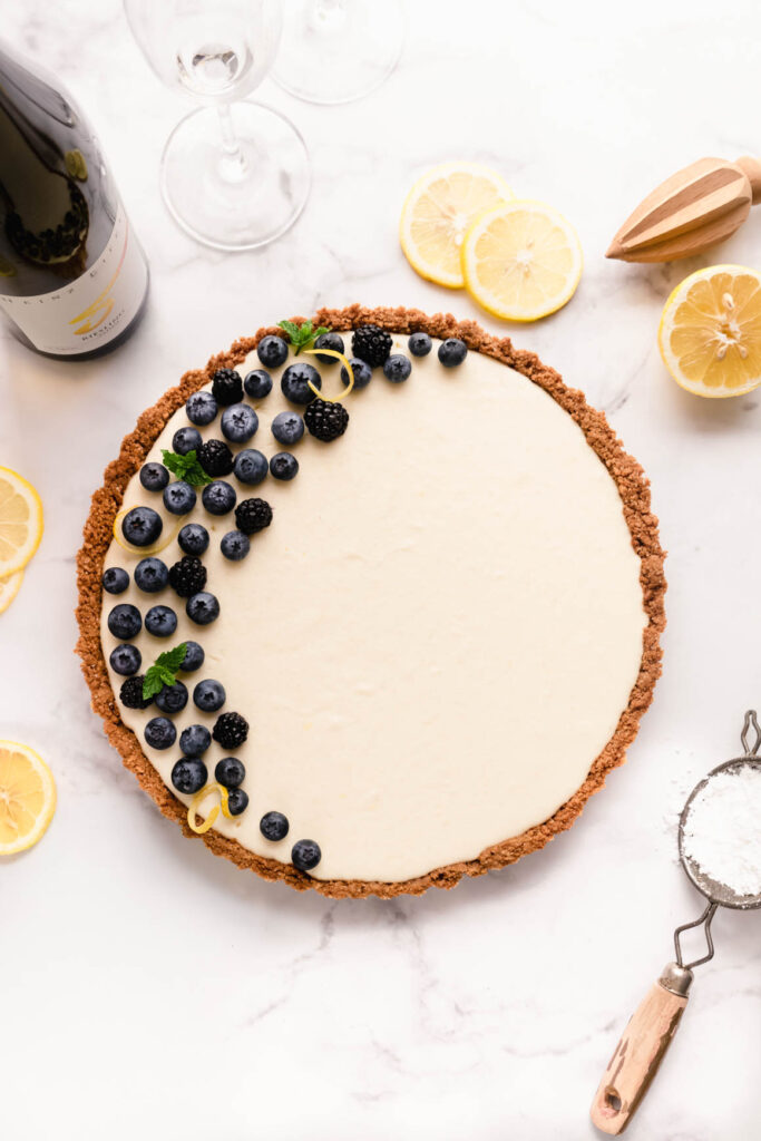 No-bake cheesecake with mascarpone in a graham cracker and gingersnap crust, topped with fresh blueberries, blackberries, lemon zest, and mint, styled overhead on a white marble surface with sliced lemons and baking tools.