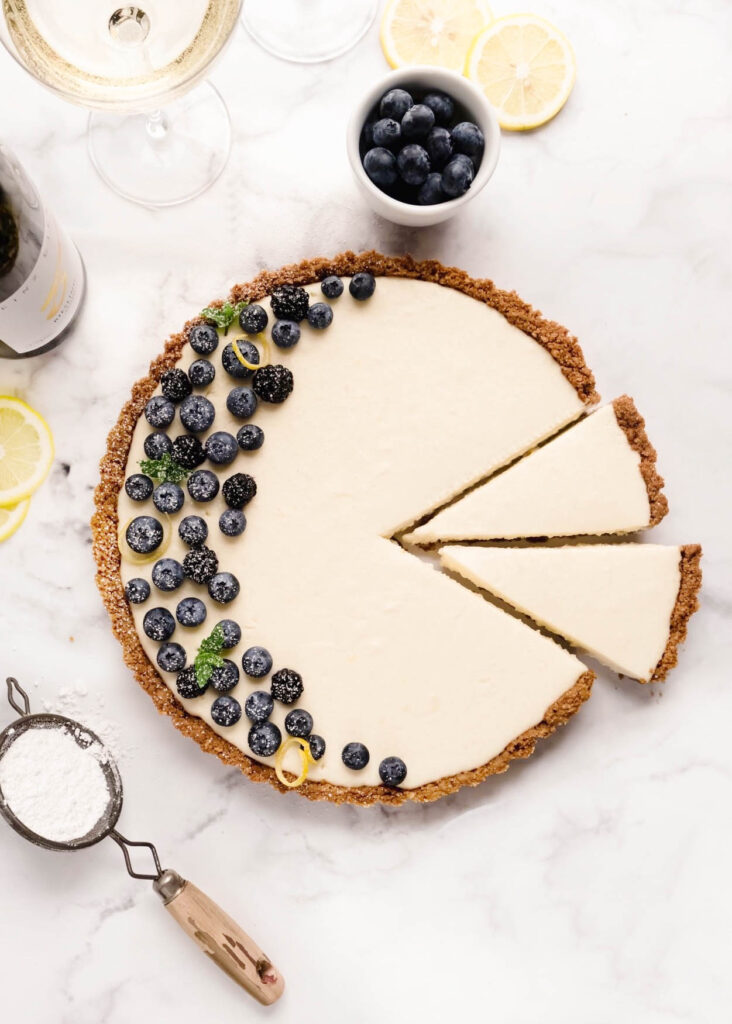 Overhead view of no-bake cheesecake with mascarpone in a graham cracker and gingersnap crust, partially sliced and topped with fresh blueberries, blackberries, lemon zest, and mint on a white marble surface.